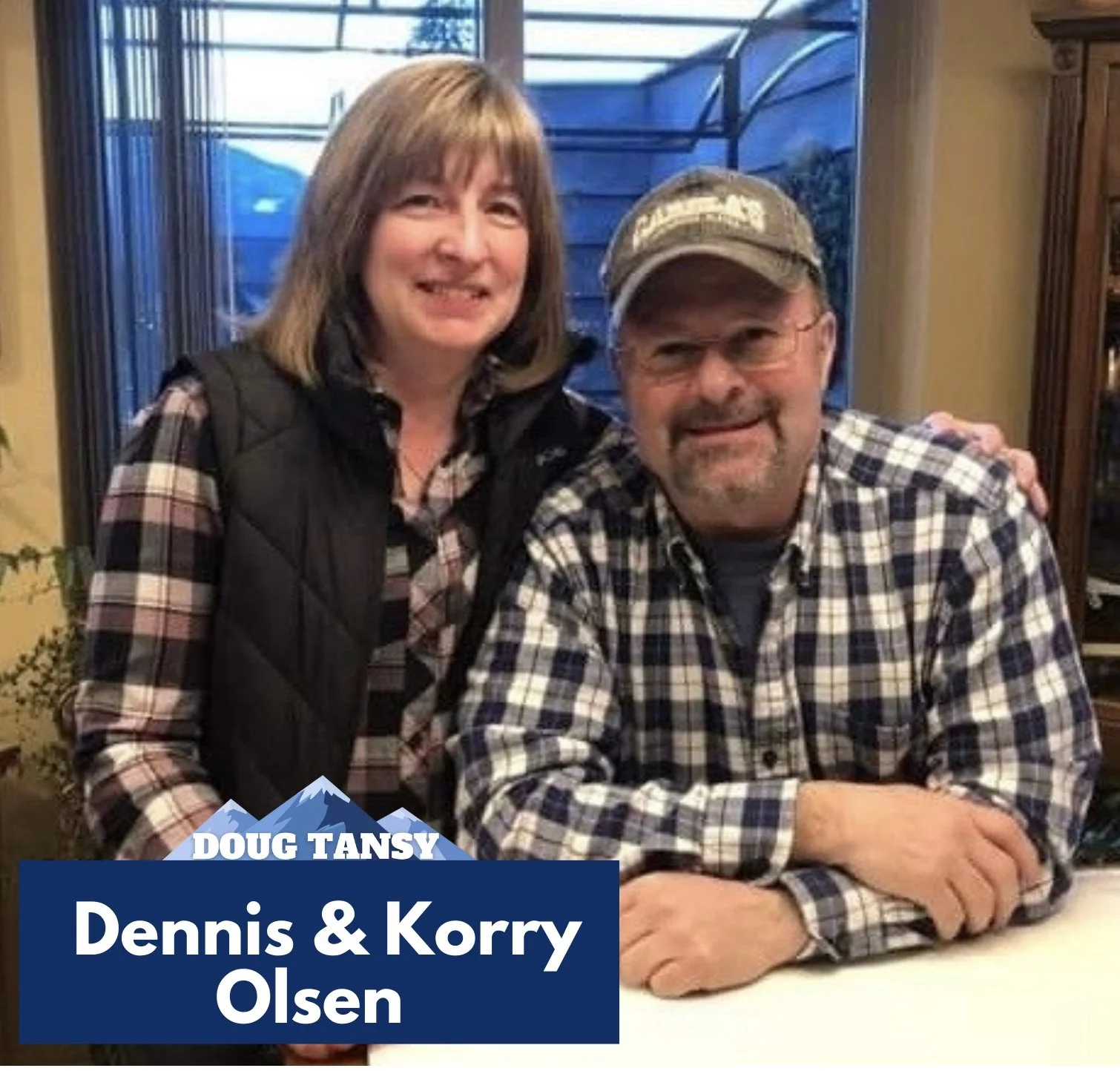 A woman and a man smiling indoors; the man is sitting at a table with his arms crossed, wearing a cap and plaid shirt, while the woman stands beside him, arm around his shoulder, wearing a plaid shirt and vest, background shows large windows and furniture. There is text overlay with mountains and the names 'Doug Tansy' and 'Dennis & Korry Olsen'.
