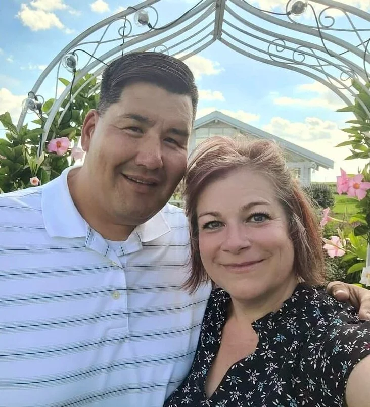 A man and woman smiling outdoors under decorative garden arch with pink flowers and greenery.
