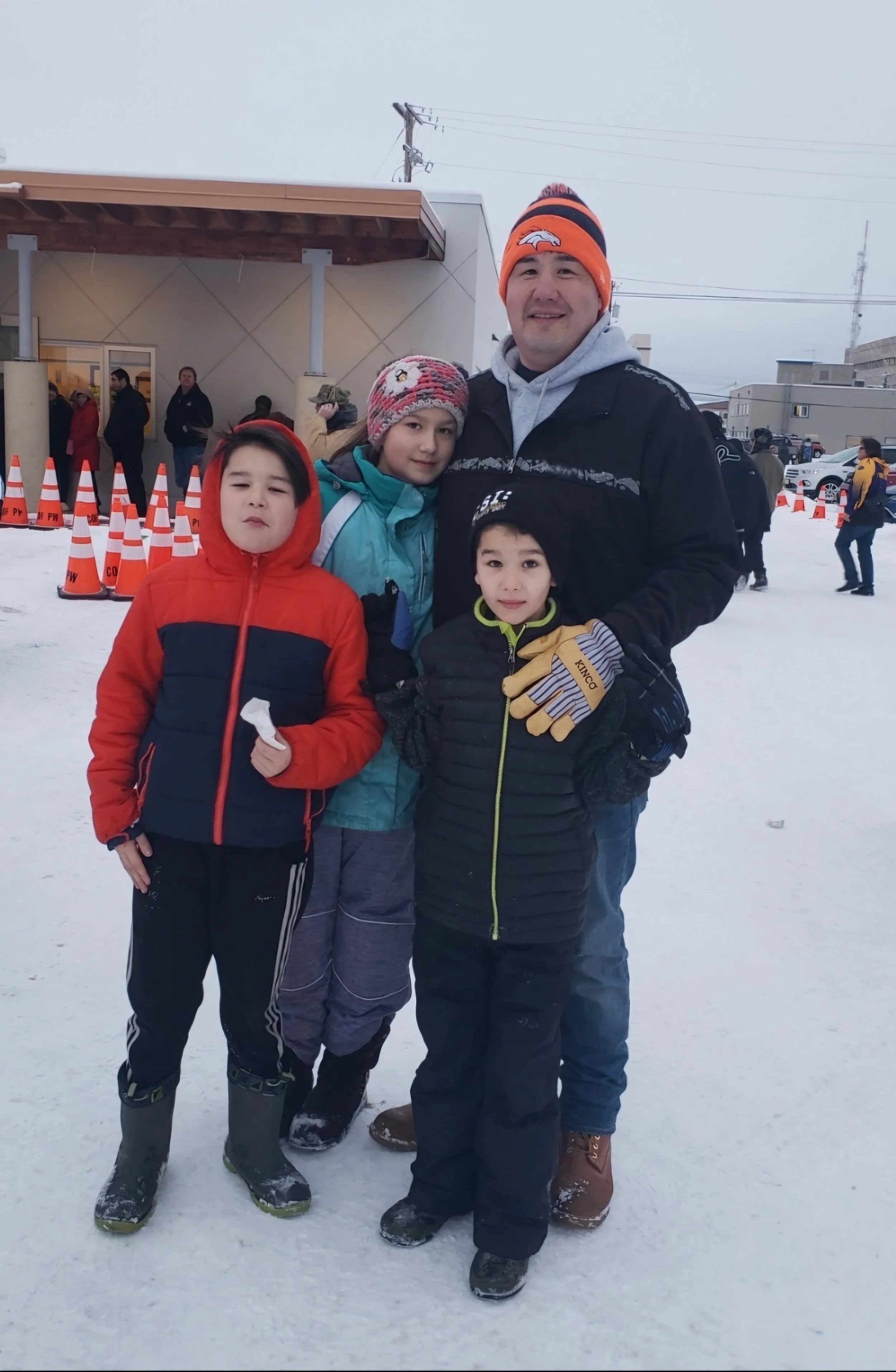 A man and three children standing outdoors in winter snow, dressed in warm clothes, with orange cones and a building in the background.