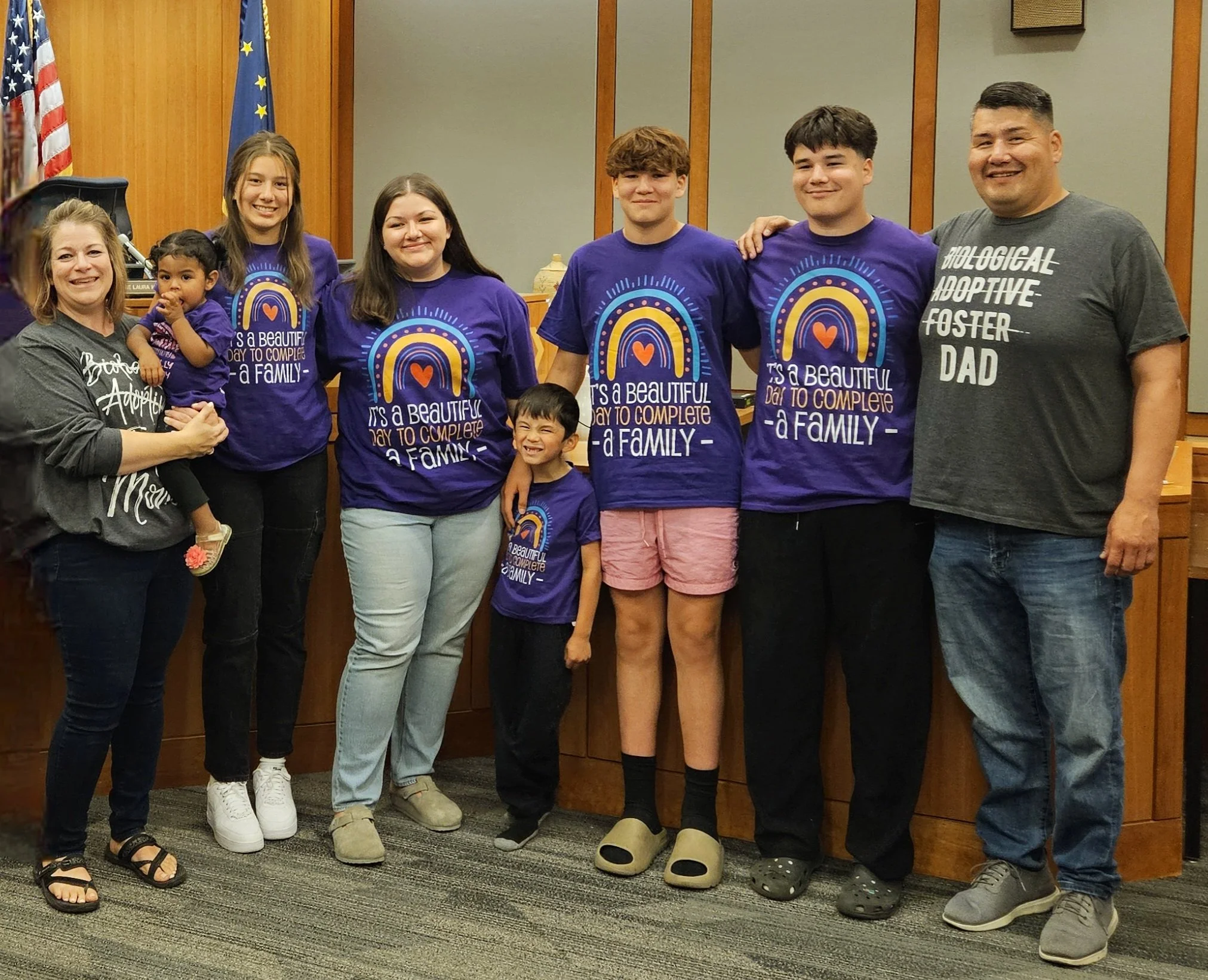 Group of eight people, including two children and six adults, standing together indoors in front of flags and wooden panels. Several individuals are wearing matching purple T-shirts with rainbow and heart designs and the phrase, 'It's a beautiful day to complete a family.' One man on the right is wearing a dark gray T-shirt that says 'Biological Adoptive Foster Dad.'