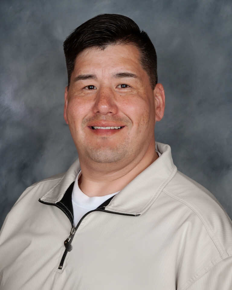 Headshot of a smiling man with short dark hair, wearing a off-white zip-up jacket and white t-shirt, posed against a grey background.