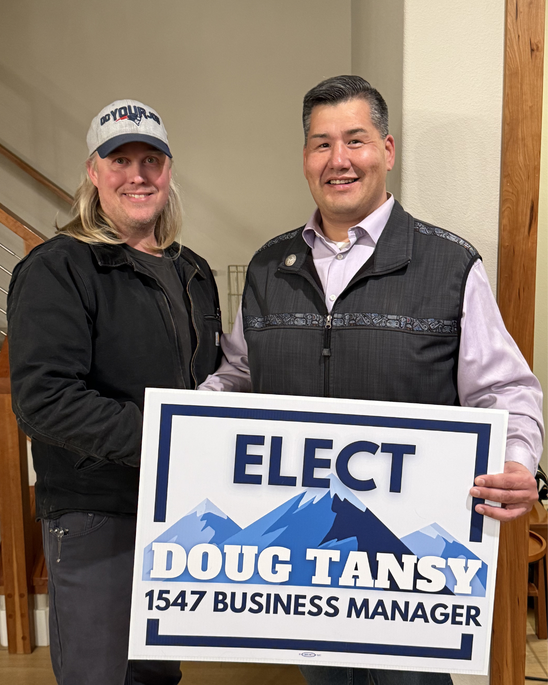 Two men standing inside, one holding a campaign sign that reads 'Elect Doug Tansy 1547 Business Manager.' The man on the left has long blond hair, glasses, and is wearing a gray baseball cap with 'Do Your Job' on it, and a black jacket. The man on the right has short dark hair and is dressed in a light pink shirt with a vest. They are smiling.