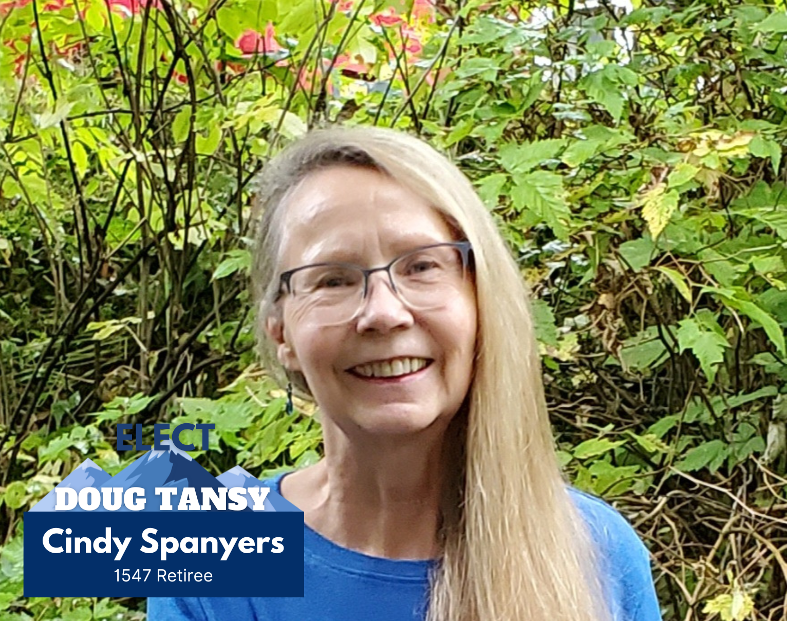 A woman with long blonde hair, glasses, and a blue shirt smiling in front of green foliage and pink flowers, with a campaign sign overlay indicating she is Cindy Spanyers, a retiree and candidate named Doug Tansy.