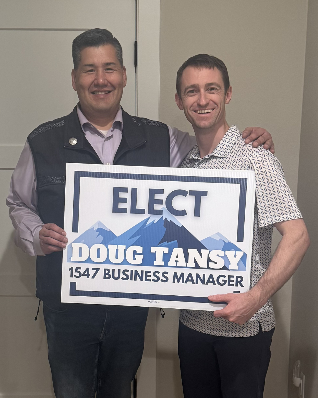 Two men smiling and holding a campaign sign that says 'Elect Doug Tansy 1547 Business Manager' with mountains in the background.