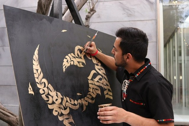 Man painting a black canvas with a design in tan, elaborate calligraphy style