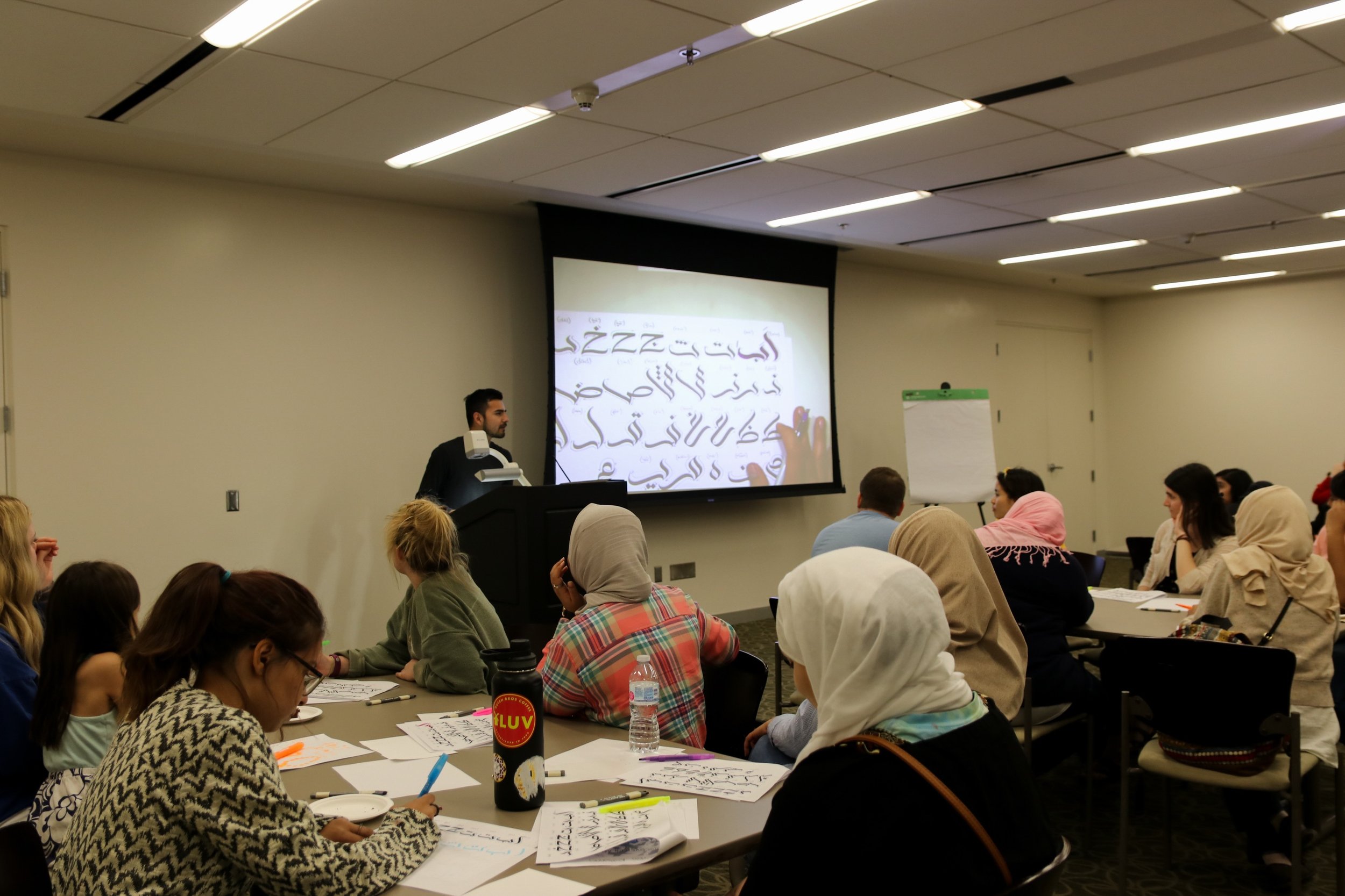 A woman in a pink headscarf and a woman in a beige headscarf sitting at a table during a workshop or class, with a male instructor at the front and a projector screen displaying Arabic script.