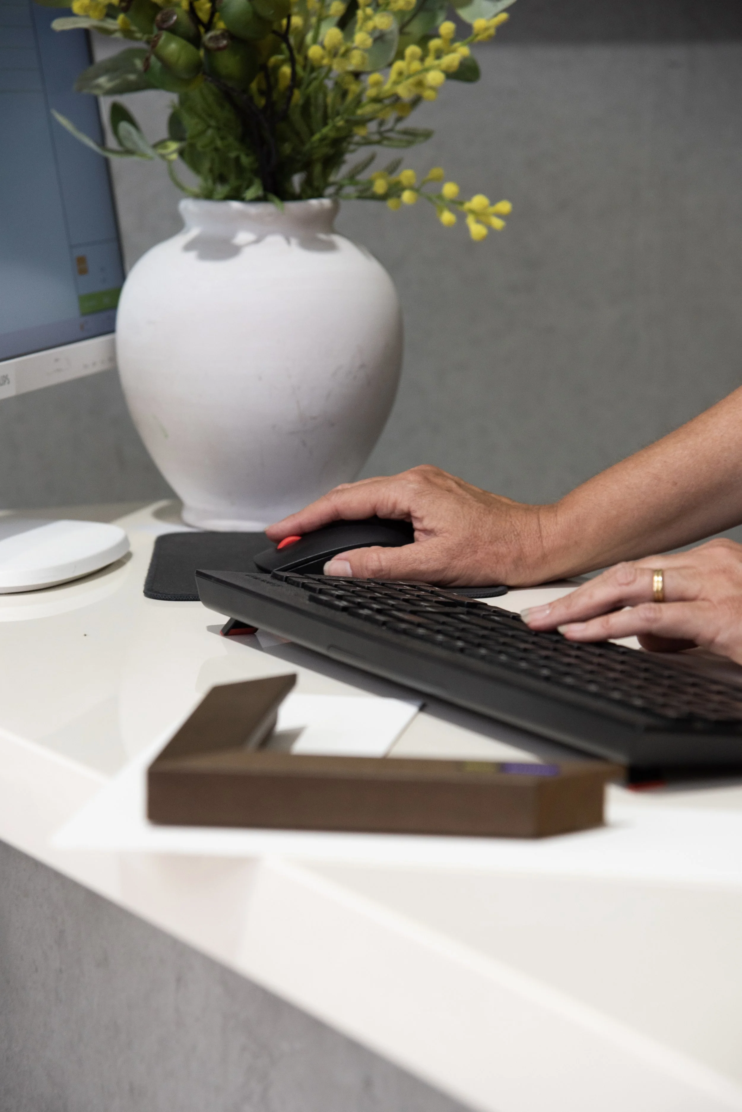 A workspace with a computer monitor, a person using a keyboard and mouse, and a white vase with yellow and green artificial flowers on a desk.