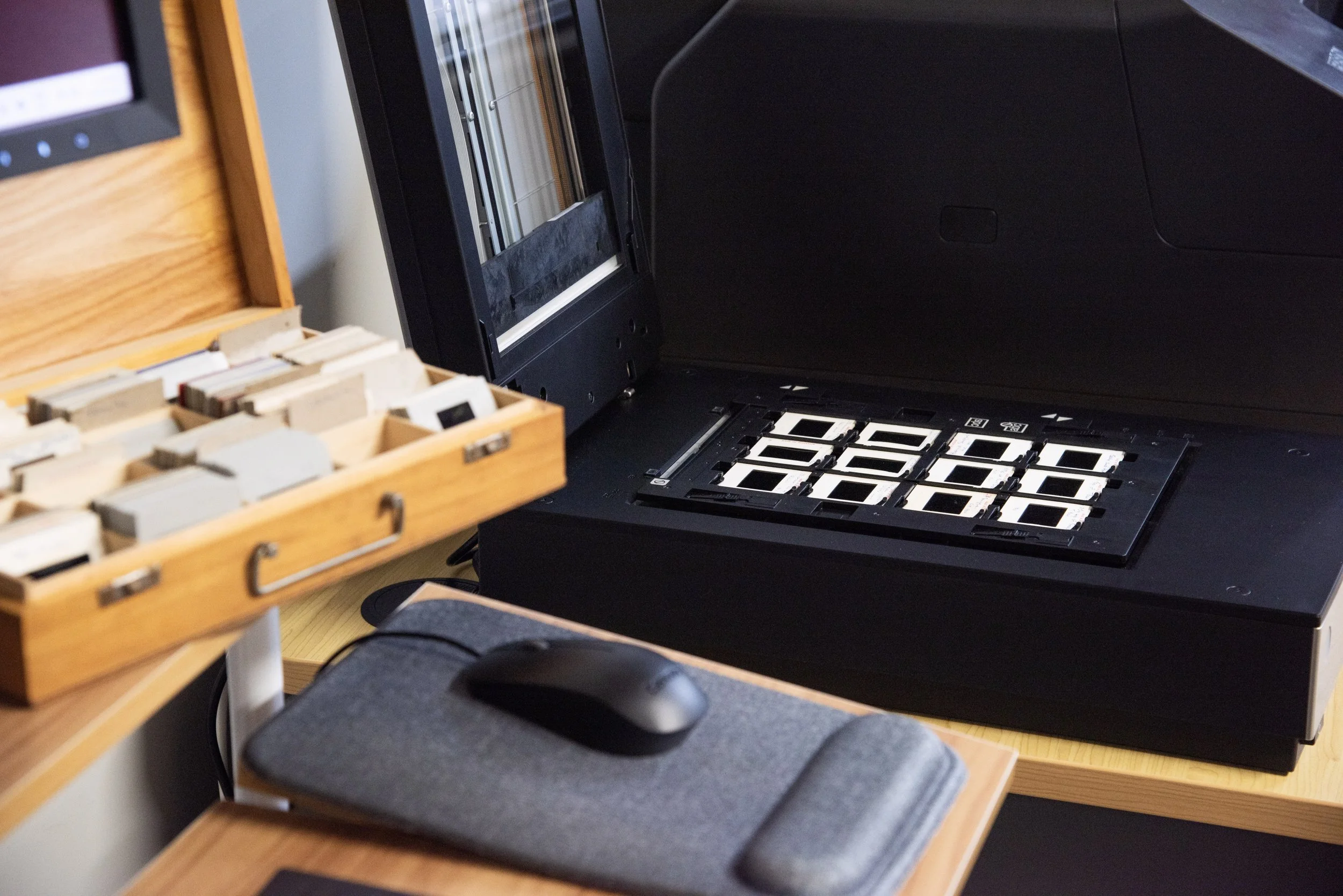 Close-up of a computer or electronic device with a keyboard or keypad, partially open wooden drawer with various small items, mouse on mouse pad, and a black plastic case or device on a wooden desk.