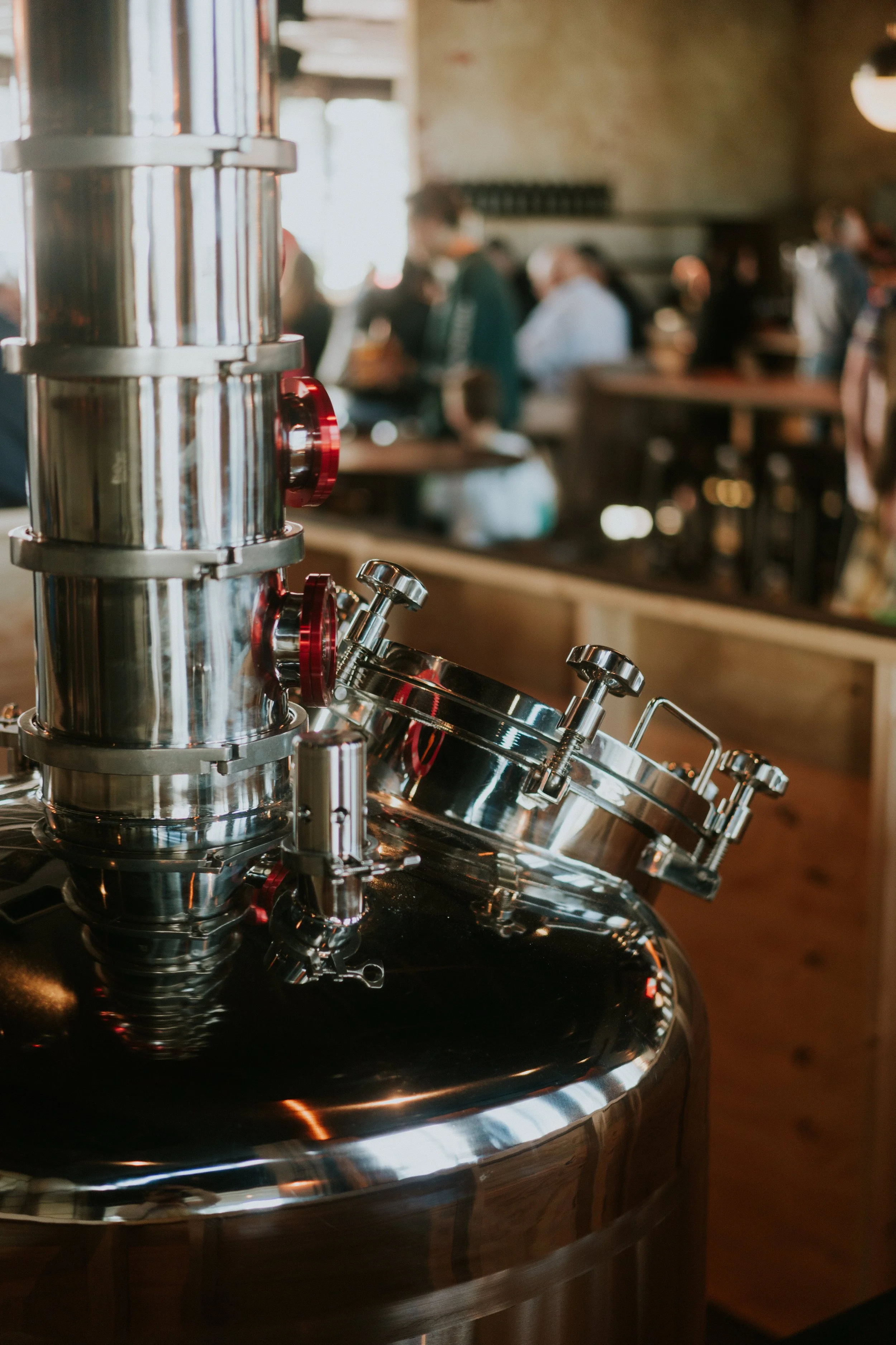 Large stainless steel brewing equipment in a brewery with people in the background.