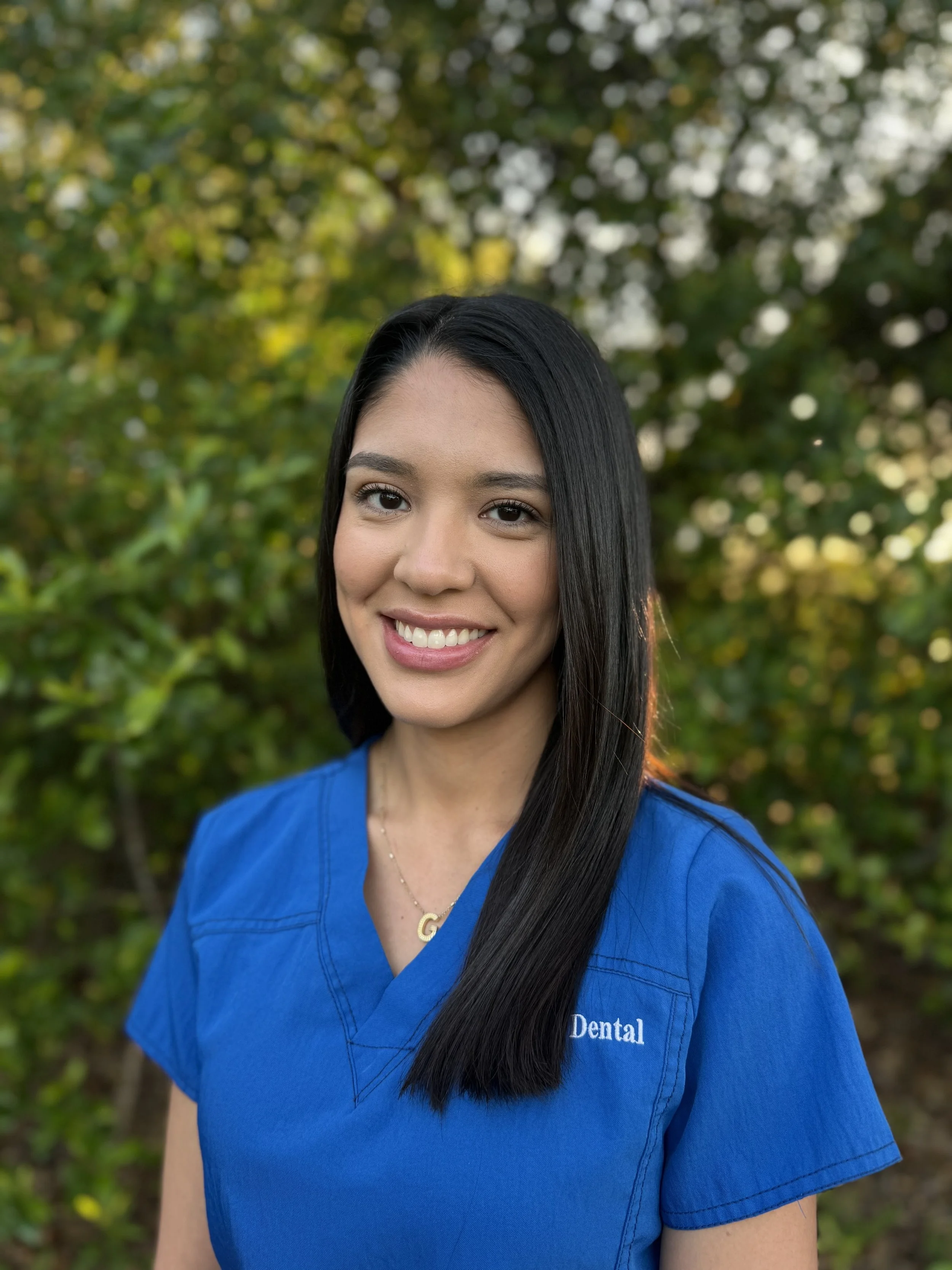 A smiling woman with long black hair wearing a blue dental uniform with the word 'Dental' embroidered on it, standing outdoors with blurred green foliage in the background.