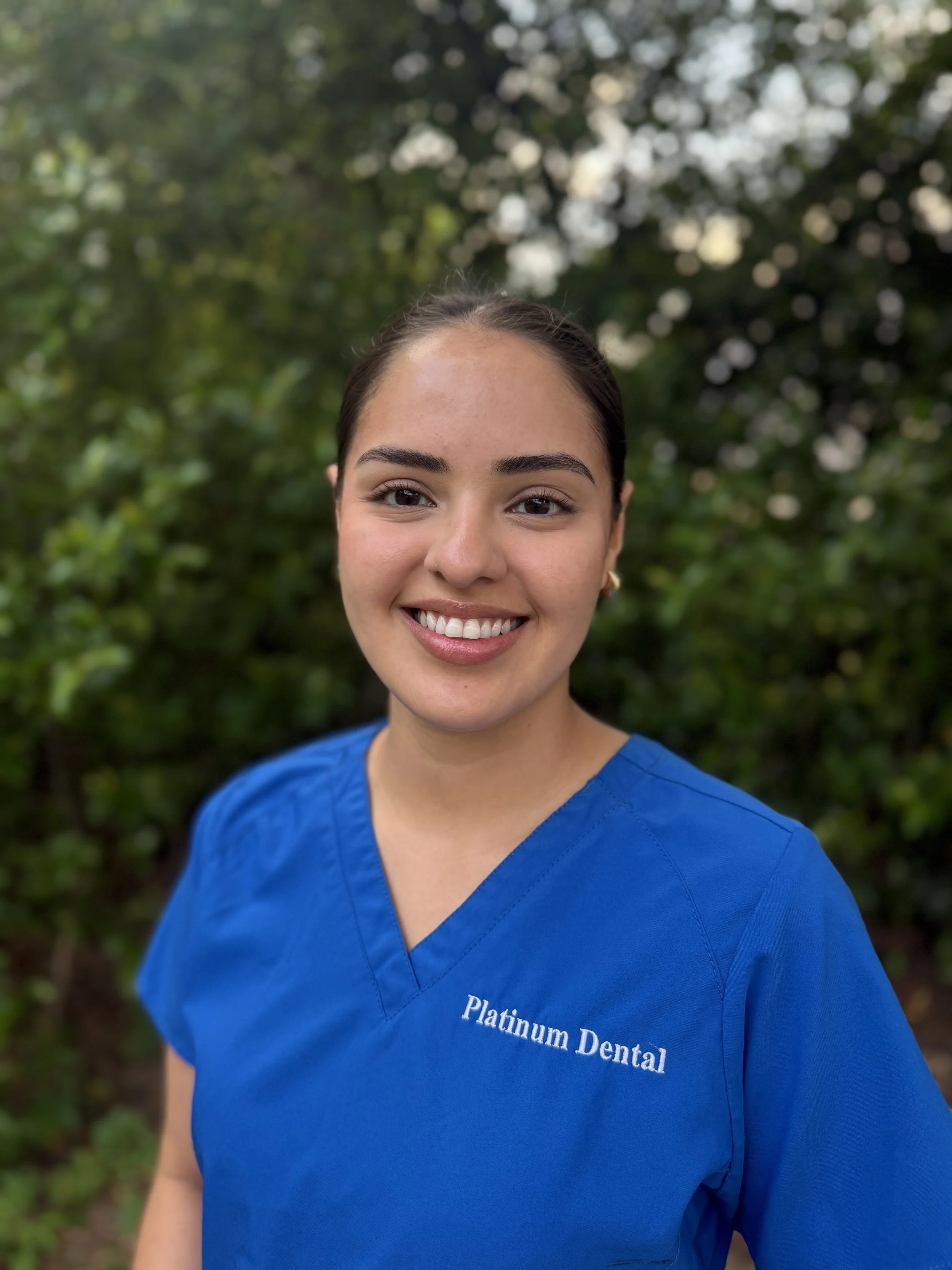 Close-up of a woman wearing a blue dental scrub top with 'Platinum Dental' embroidered on it, smiling outdoors with a background of green trees.