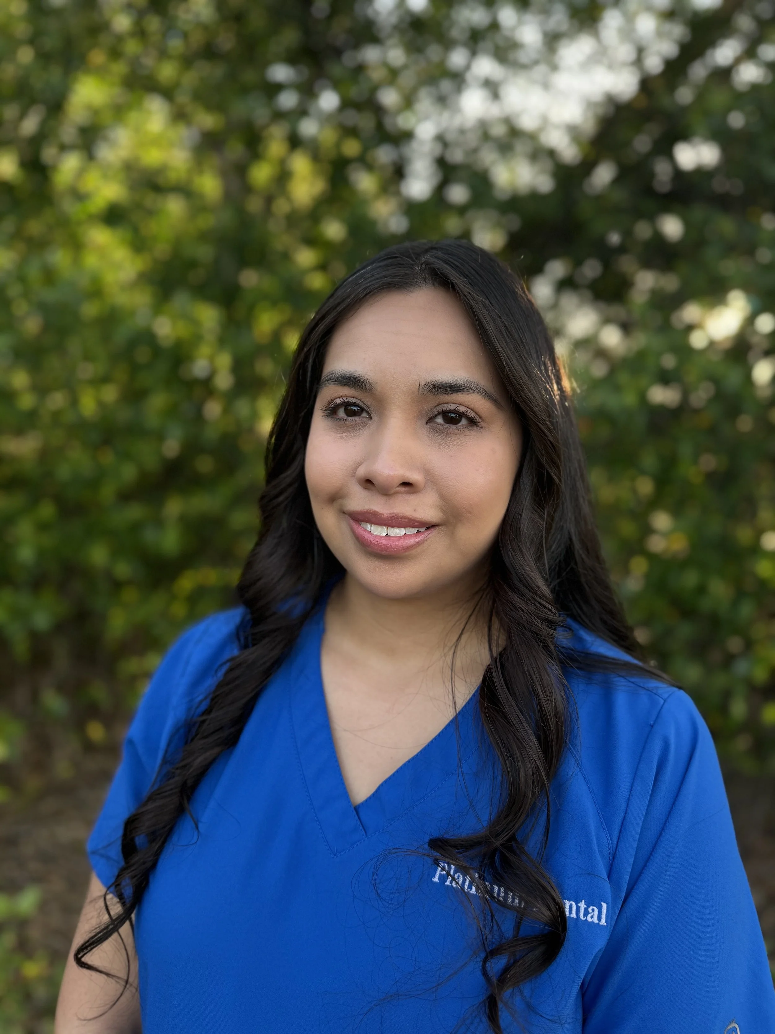 A woman with long black hair wearing a blue medical uniform, standing outdoors with blurred green trees in the background.