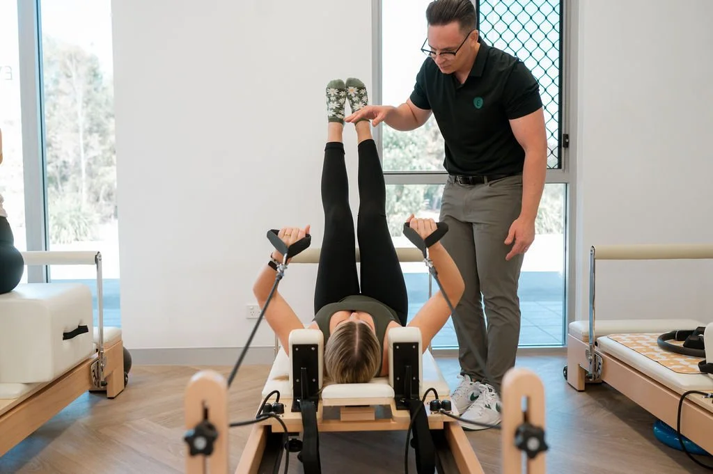 A woman undergoing physical therapy on a reformer machine while a therapist assists her, in a well-lit room with large windows.
