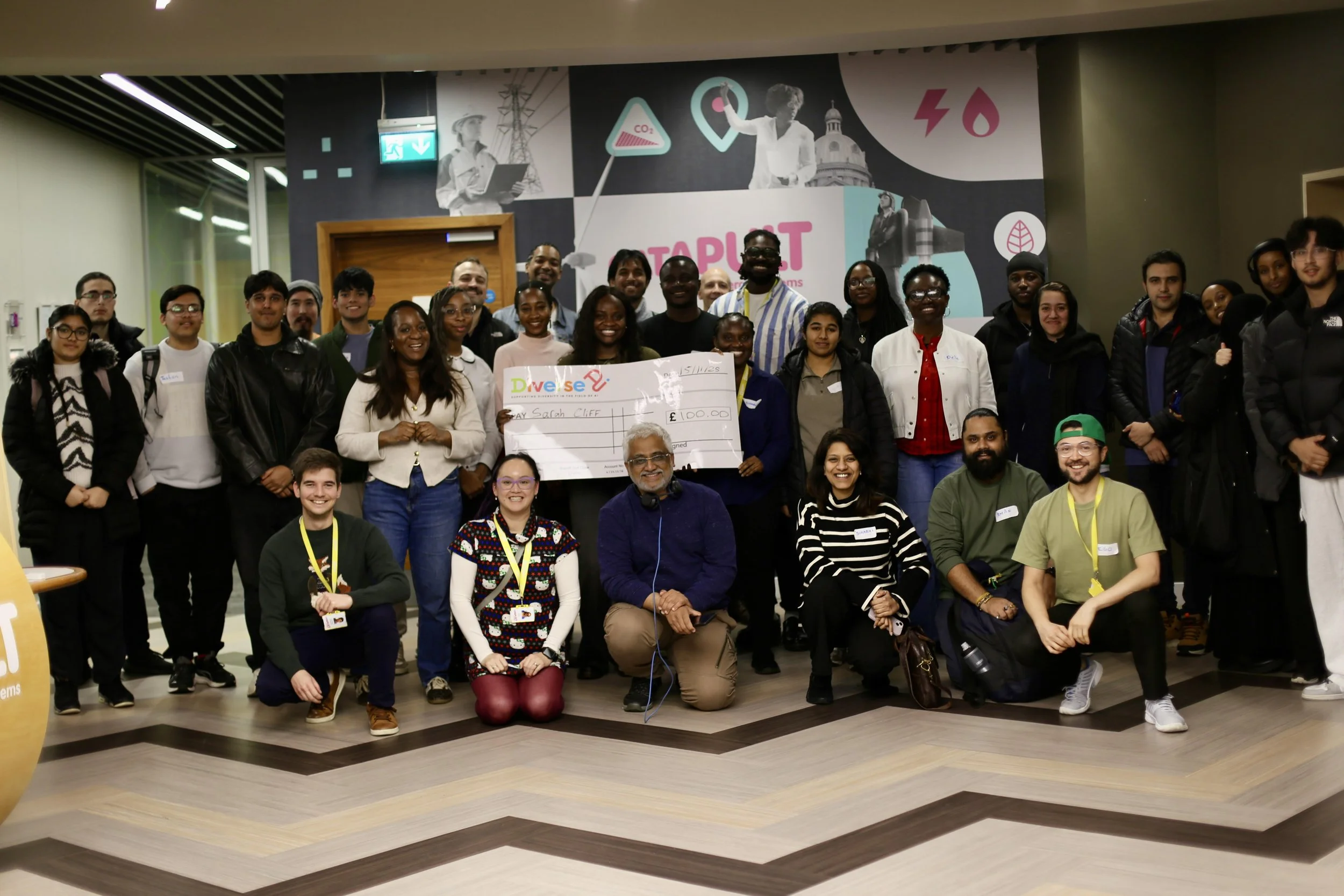 A large group of diverse people gathered indoors, smiling for a group photo. Several individuals are holding a large check for 100 pounds, indicating a donation or award event.