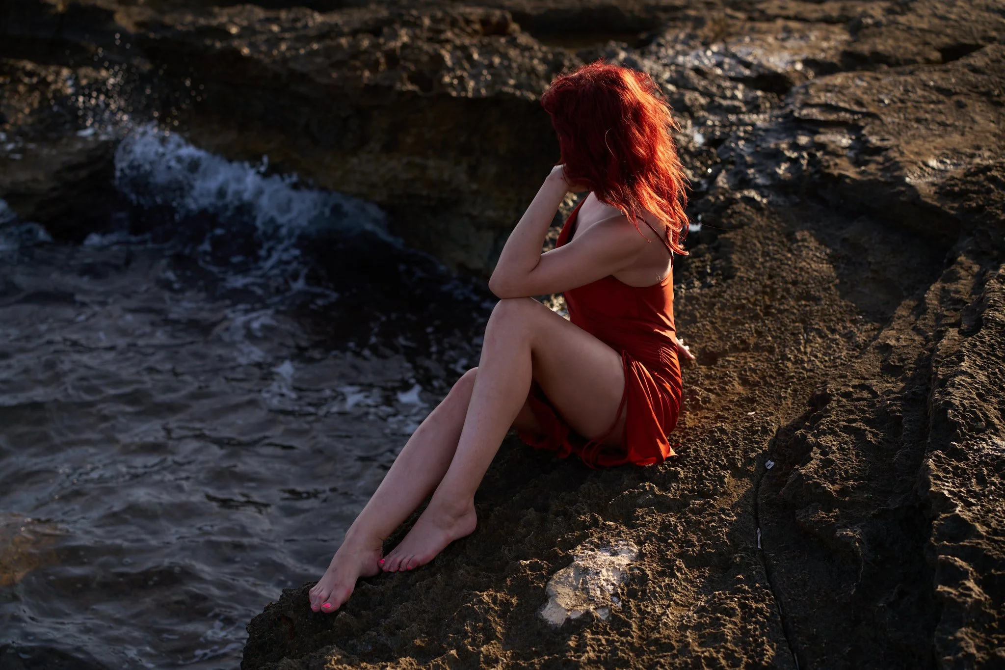 Woman with red hair in a red dress sitting on rocky shoreline near water