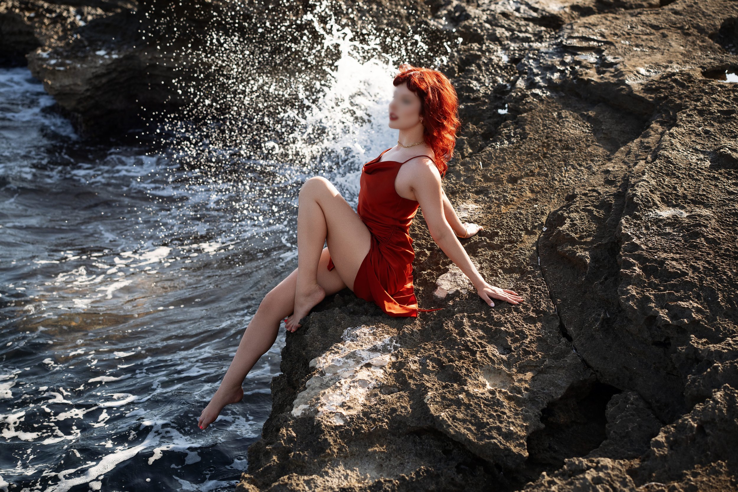 Woman with red hair in a red dress sitting on rocks by the ocean, with waves splashing nearby.
