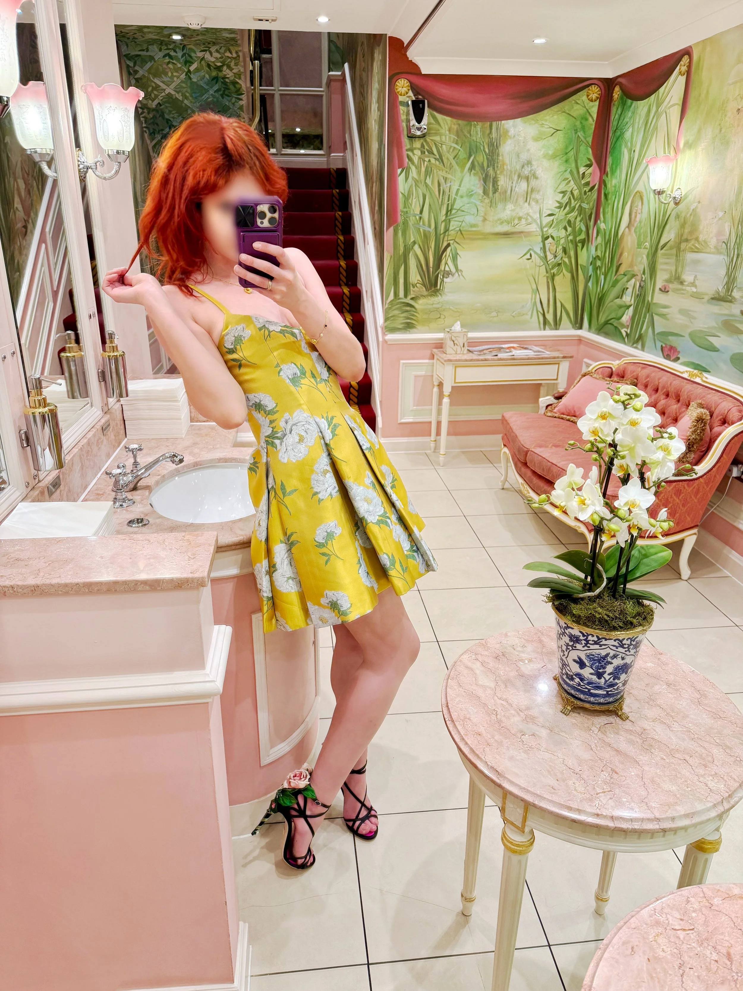 A woman with red hair taking a mirror selfie in a pink-themed, elegant room with a mural of nature on the wall, a pink sofa, a small side table, and a potted plant on a marble-topped table.
