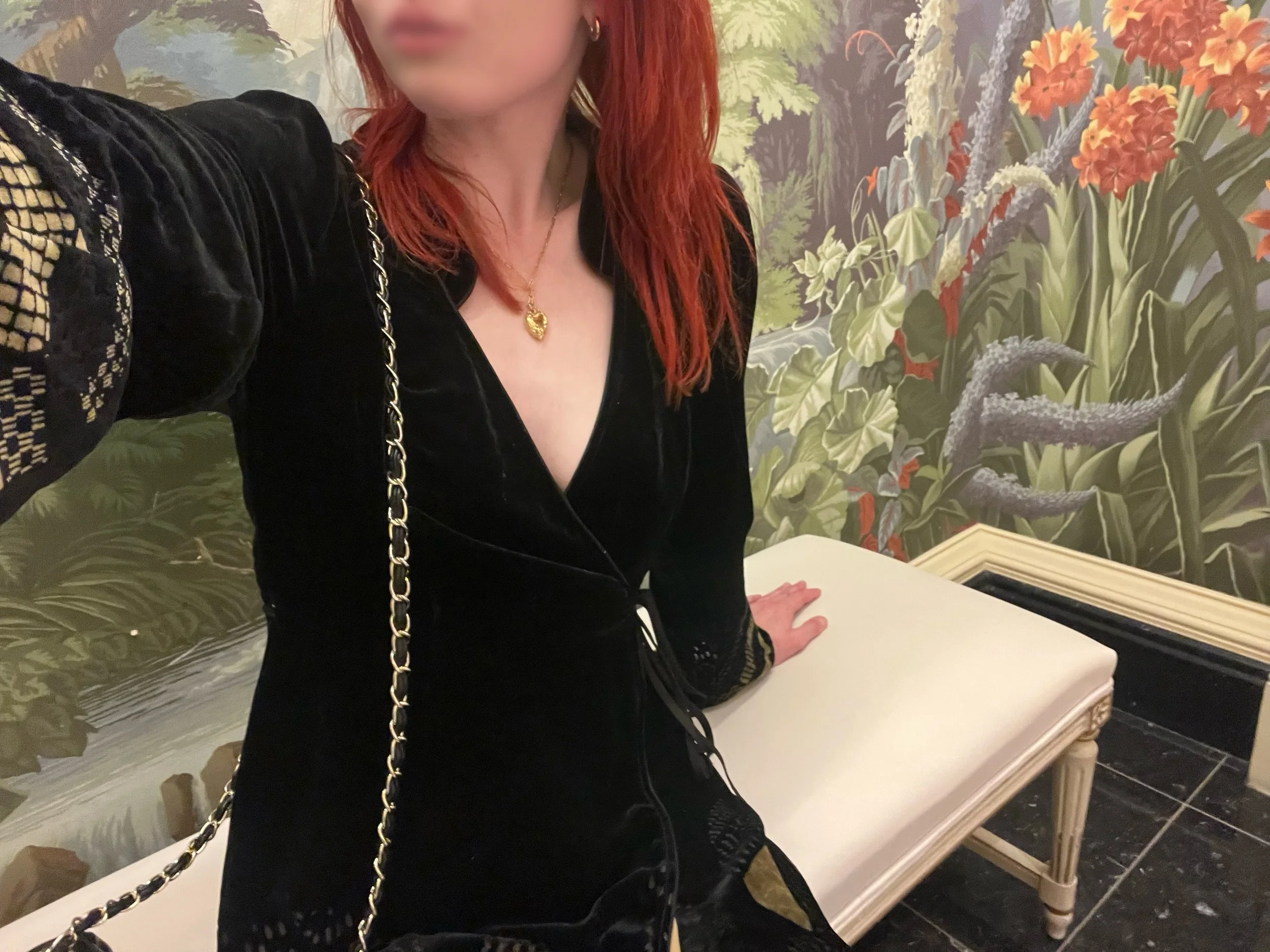 A woman with red hair taking a selfie, wearing a black dress with lace details on the sleeves and a gold chain strap purse. She is seated on a white bench in a room with floral wallpaper featuring various green plants and orange flowers.