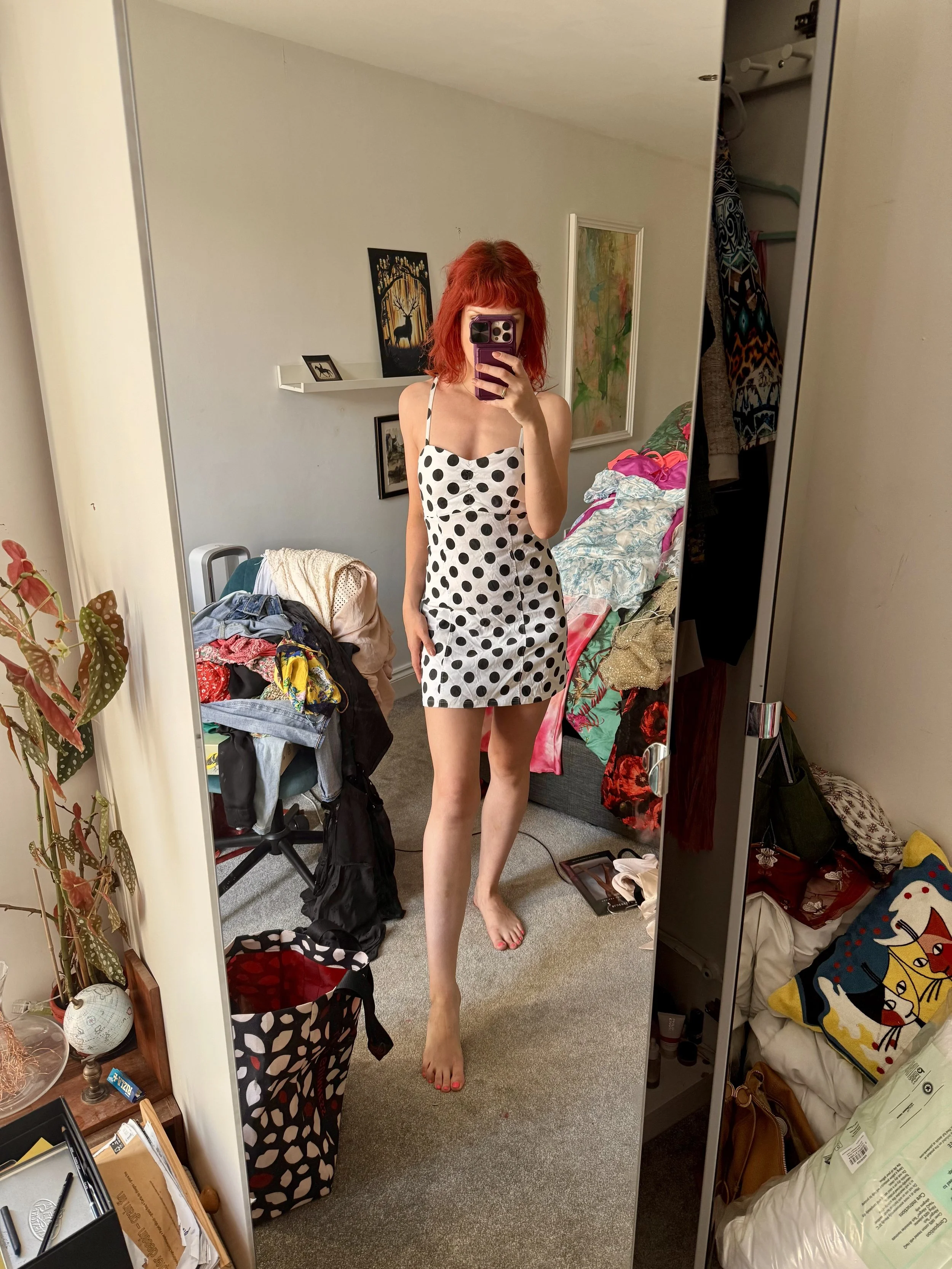 A woman with red hair taking a mirror selfie in a cluttered bedroom, wearing a black and white polka dot dress.