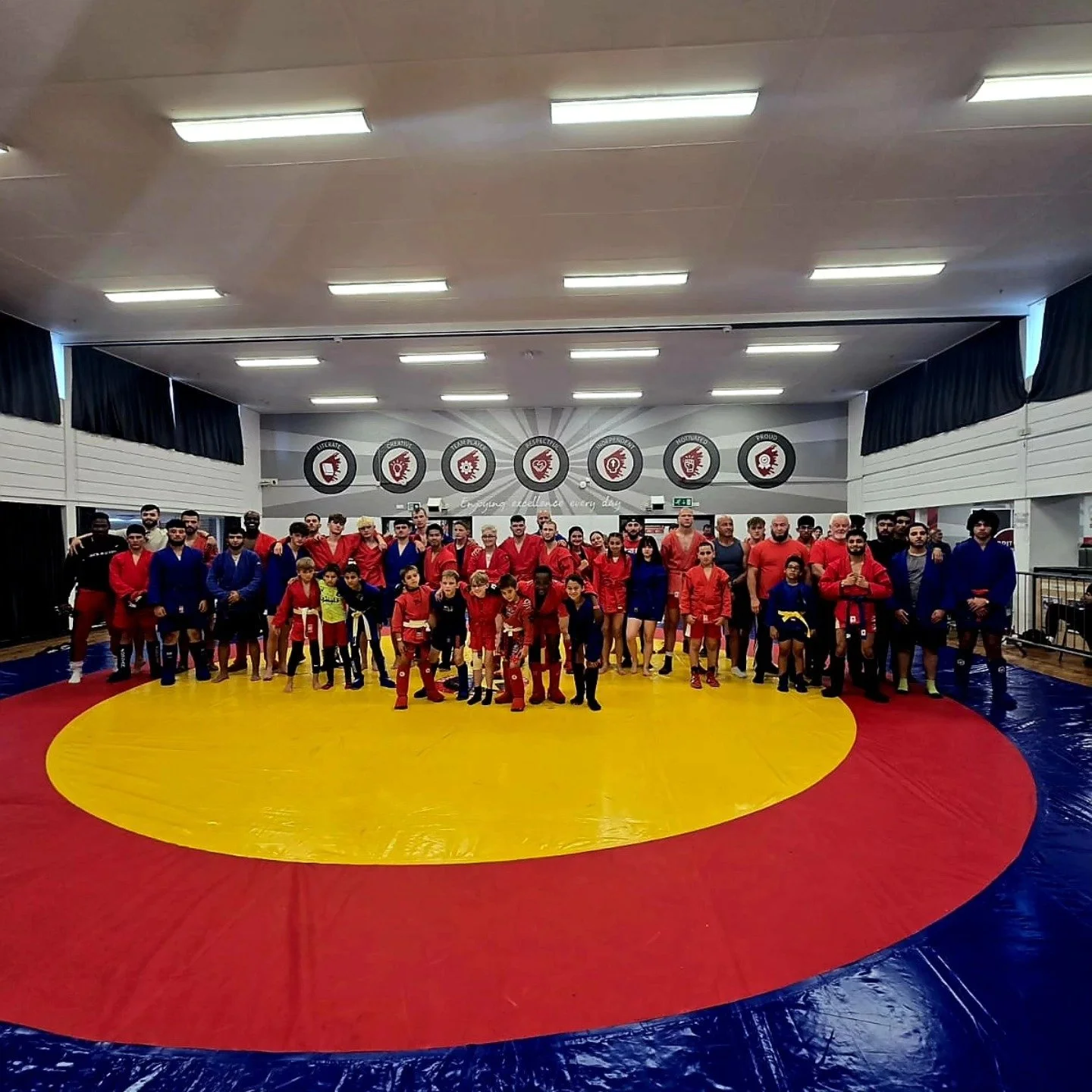 Group of martial artists and children posing on a wrestling mat inside a gym with motivational posters on the wall.