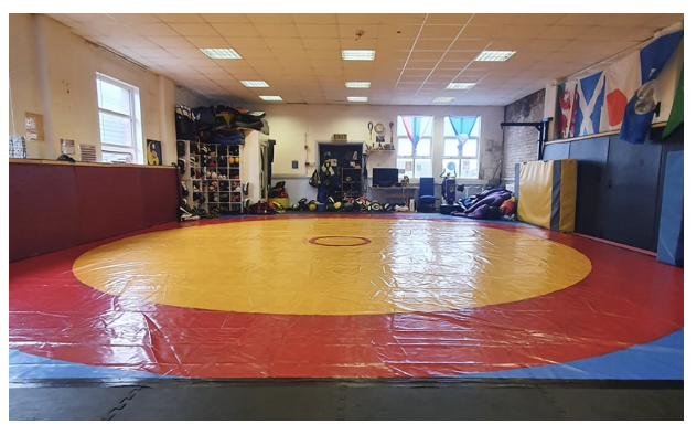 A wrestling training room with a large red and yellow mat, shelves filled with gear, and flags hanging on the wall.