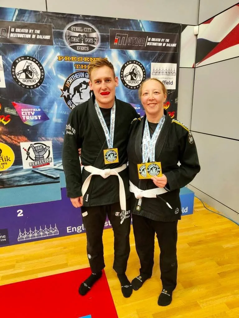Two martial artists in black gi uniforms with white belts posing after a competition, wearing medals around their necks, in front of a competition backdrop.