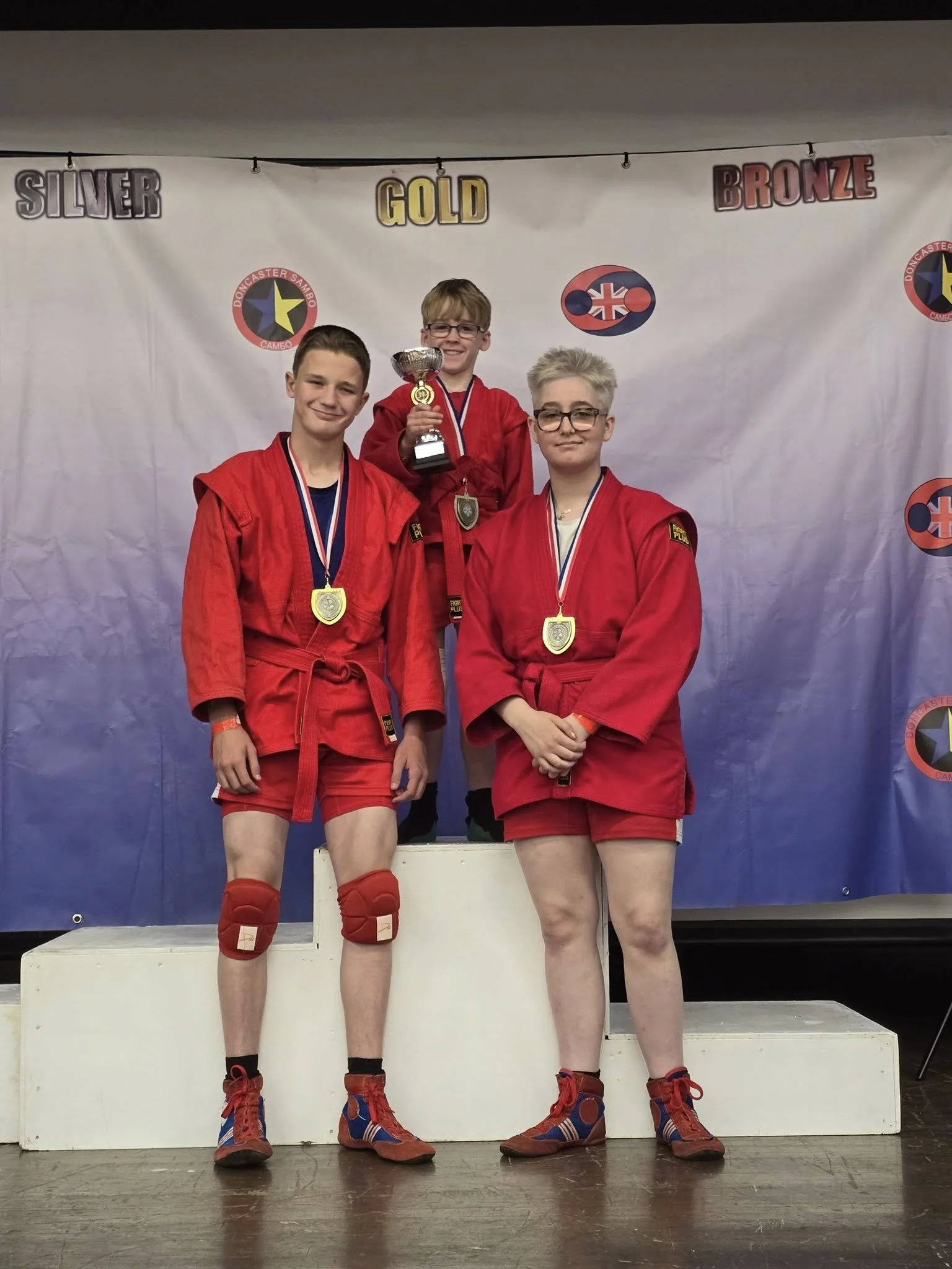 Three young athletes in red judo uniforms and medals standing on a winner's podium, with one holding a trophy, in front of a backdrop with medals labeled Silver, Gold, and Bronze.
