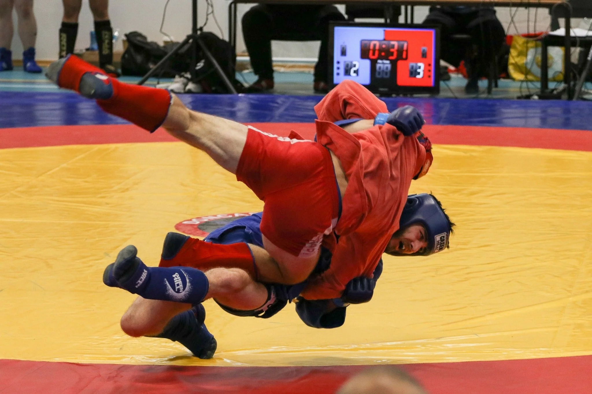Two male wrestlers in red and blue uniforms engaged in a wrestling match on a yellow mat with a red border. One wrestler is on top of the other, who is on the ground. A scoreboard in the background shows a score of 2 to 3 and 33 seconds remaining.