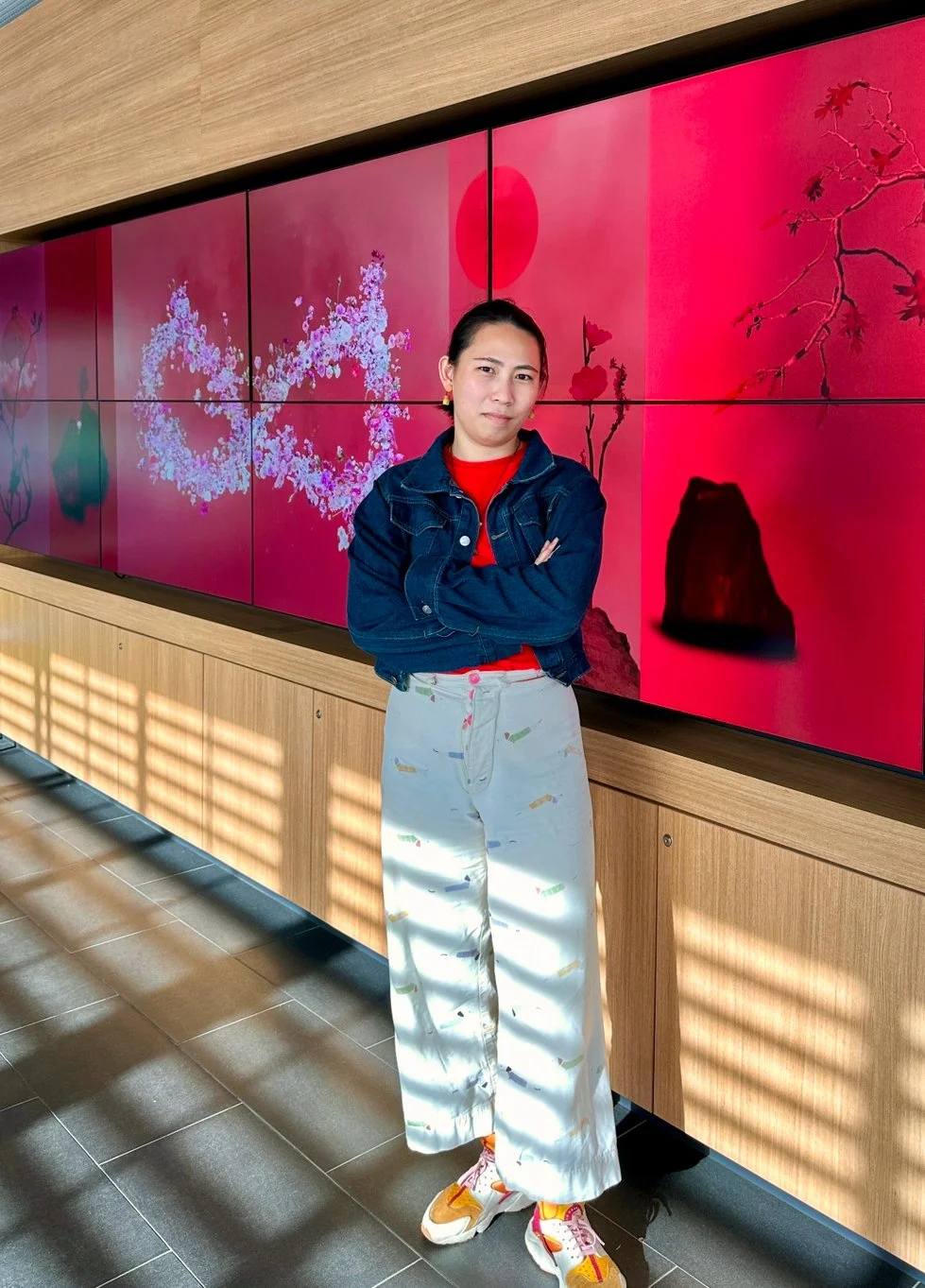 A woman stands with arms crossed in front of a digital art display with pink floral designs and a red circle, wearing a denim jacket, red shirt, light-colored patterned pants, and sneakers.