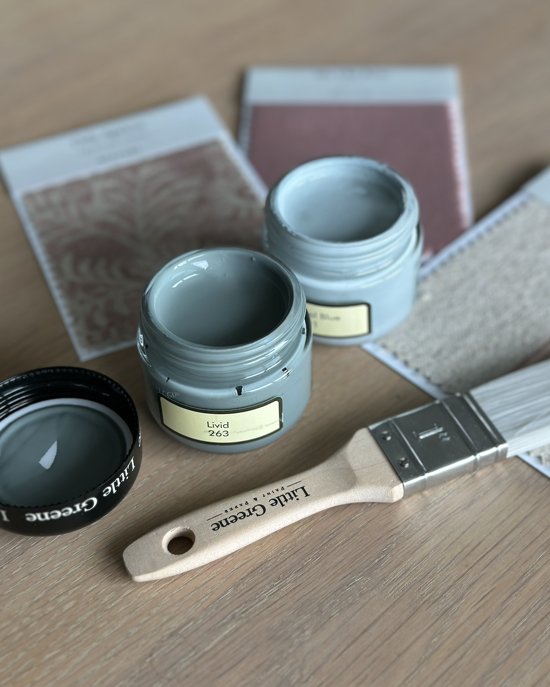 Paint supplies on a wooden surface, including two open jars of blue-green paint, a paintbrush labeled 'Lime Green', and color samples or swatches.