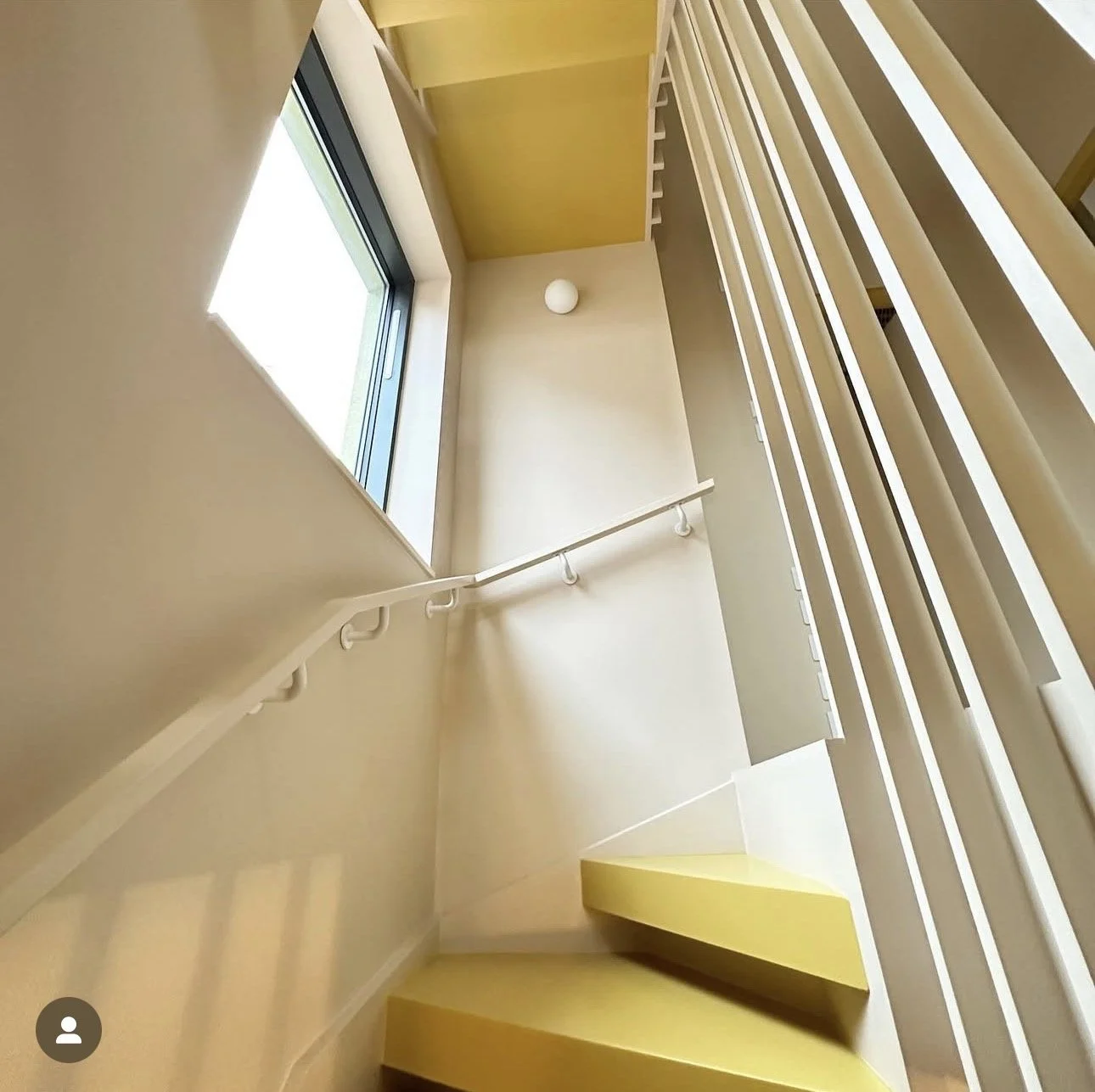 Interior view of a stairwell with a yellow wall, a large window, a white handrail, and a small yellow triangular bench at the bottom, looking upward.