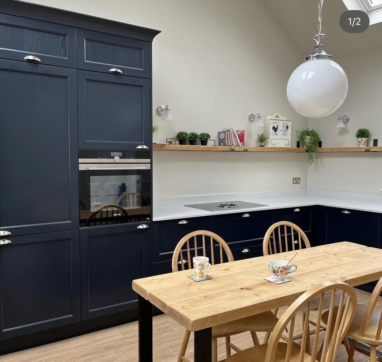 A kitchen with dark blue cabinets, a wooden table with chairs, and a white countertop. There are cups on the table, a hanging light fixture, and decorative plants and books on a floating shelf.