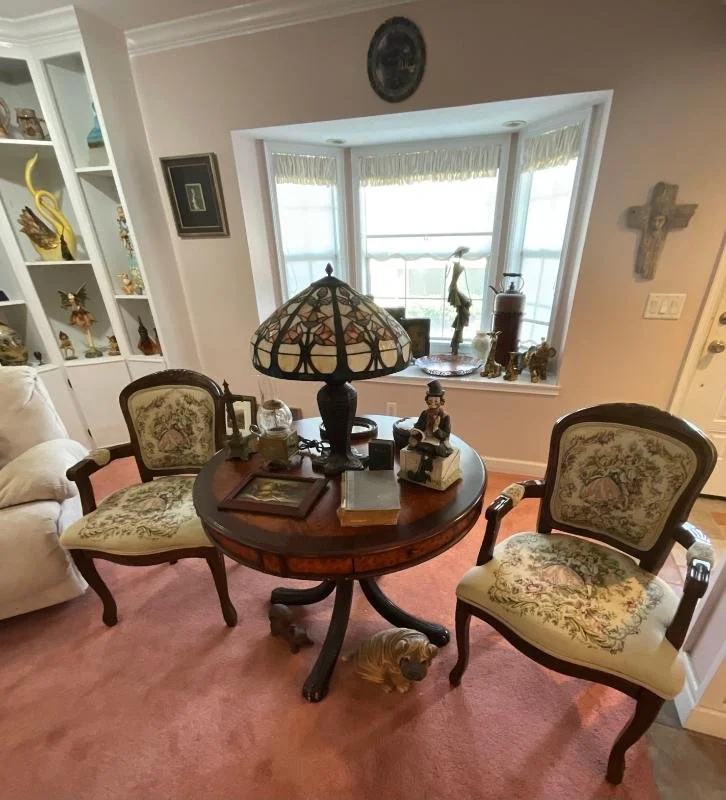 A round wooden table with decorative items and two upholstered chairs with floral patterns in a cozy living room. A stained glass lamp is on the table. In the background, a bay window with decorative items on the sill, a cross on the wall, and a built-in bookshelf with decorative objects are visible.