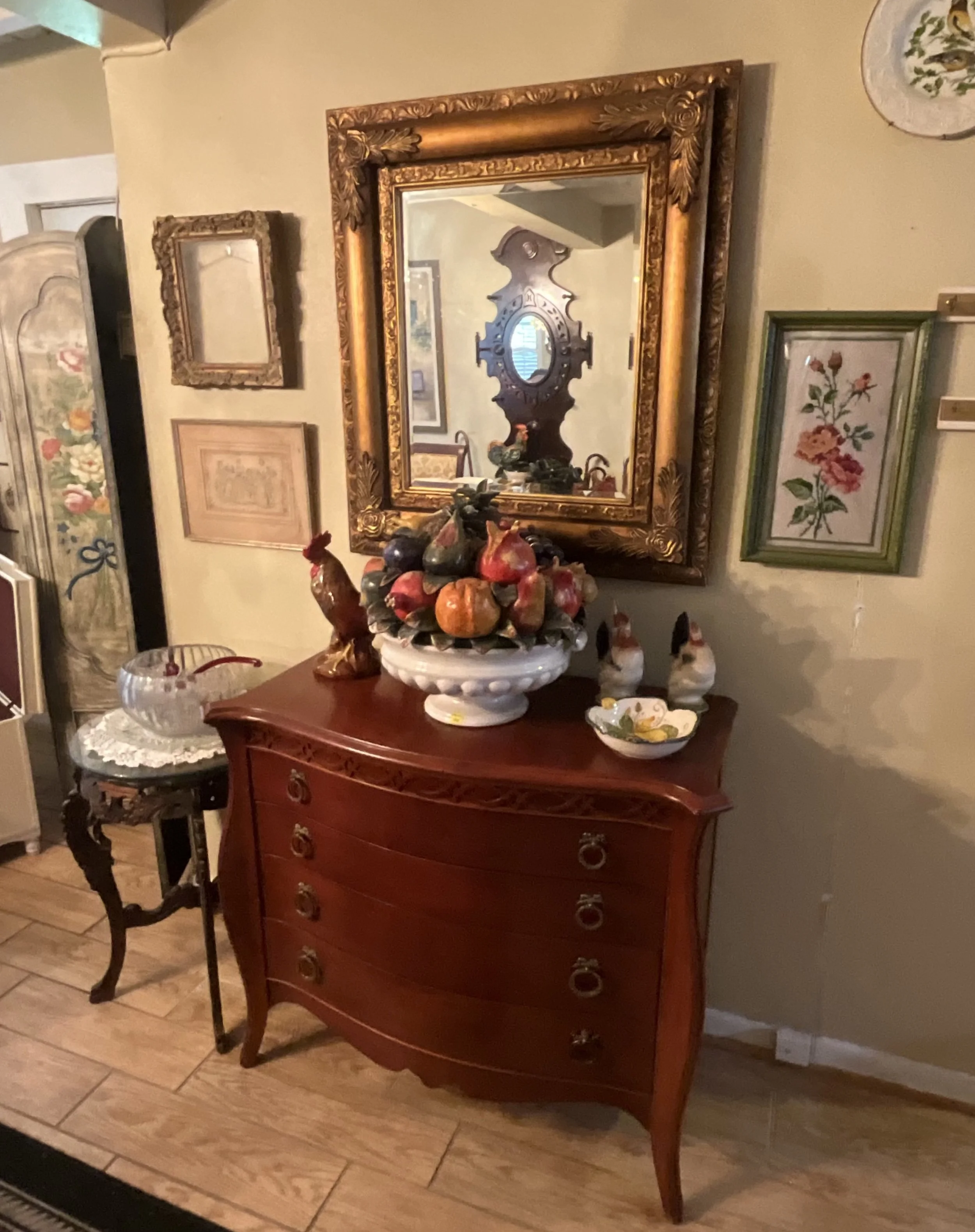 An antique wooden dresser with curved legs and brass ring pulls, decorated with a large ceramic bowl filled with faux fruit and small ceramic roosters. Above the dresser, a gold ornate mirror reflects part of the room. To the left, a small side table with a knitted doily and glass candy jar. Several framed pictures and a decorative plate hang on the wall.