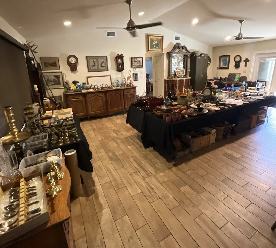 A room with wooden floors and tables covered with various collectibles and antiques. The walls have framed pictures and clocks, and there are cabinets and shelves displaying more items. The room appears to be set up for a sale or exhibition of antiques.