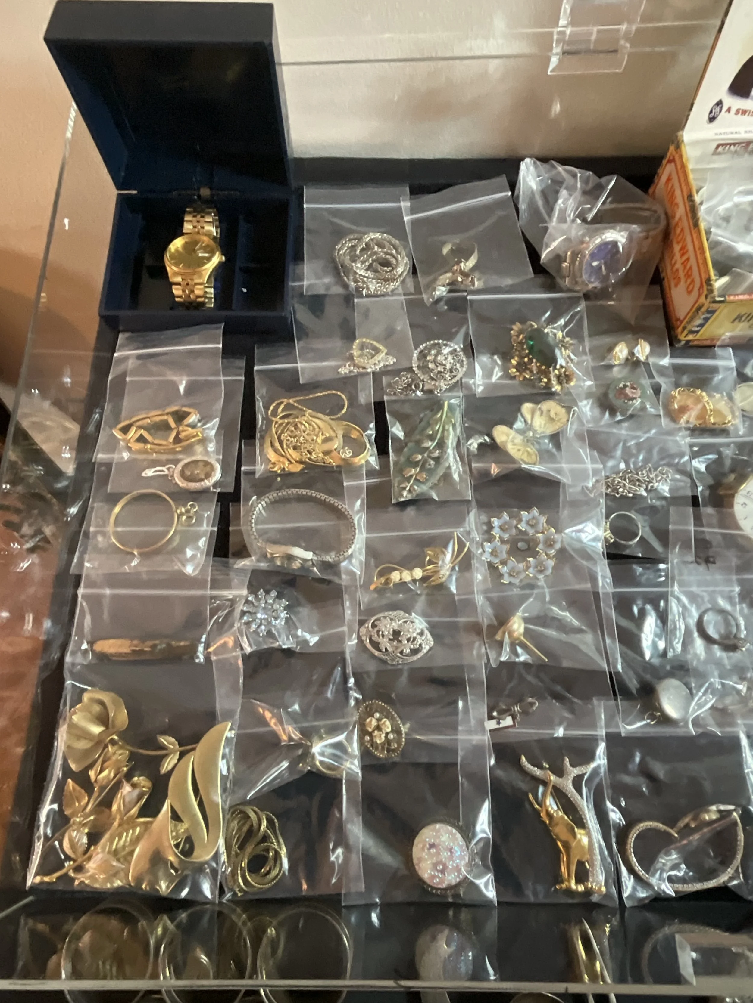 A collection of various jewelry items including rings, necklaces, brooches, earrings, and watches displayed on a black surface with some items in small plastic bags and a watch box.