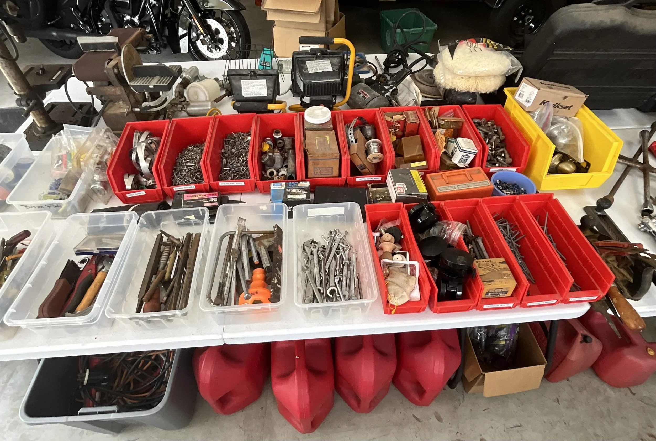 A garage workbench with organized plastic bins filled with nuts, bolts, screws, tools, and hardware, with some larger containers and power tools in the background.