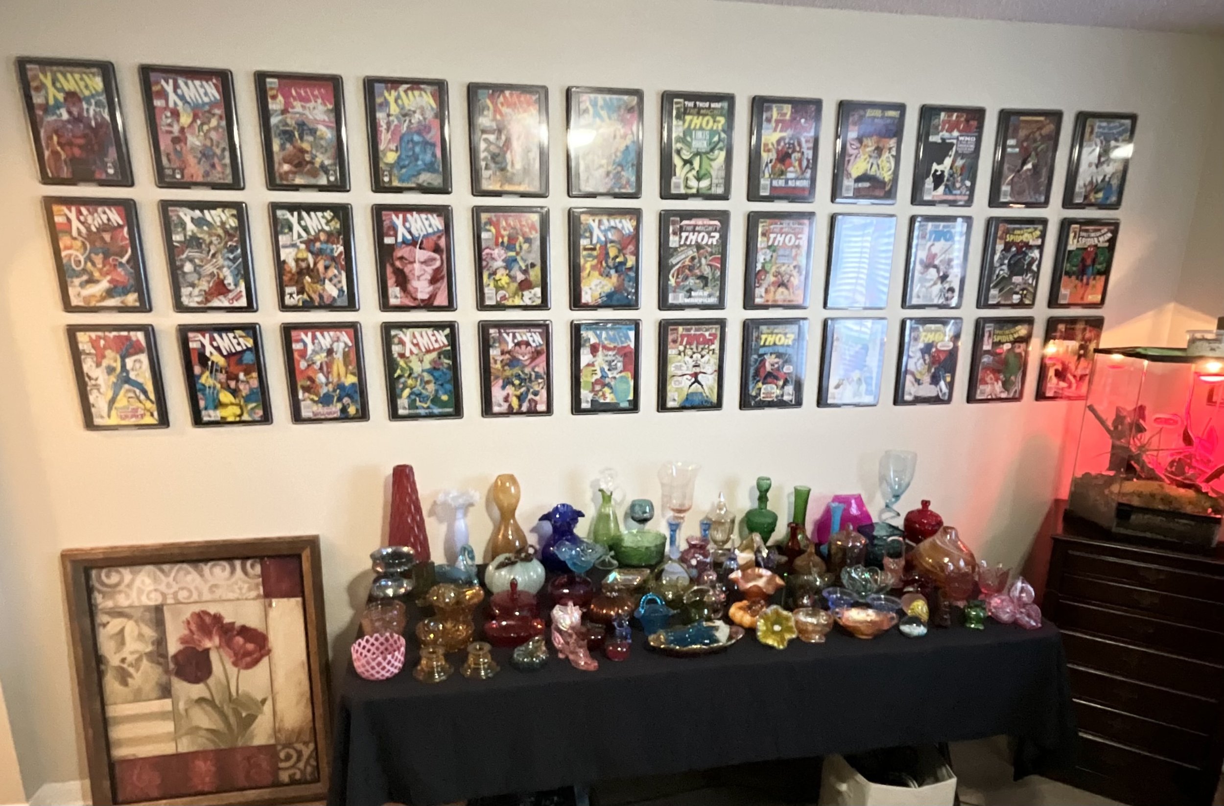 A wall decorated with framed comic book covers, with a table below filled with assorted colorful glass vases and ornaments, and a framed picture of red flowers leaning against the wall. There is a red-lit aquarium in the right corner.