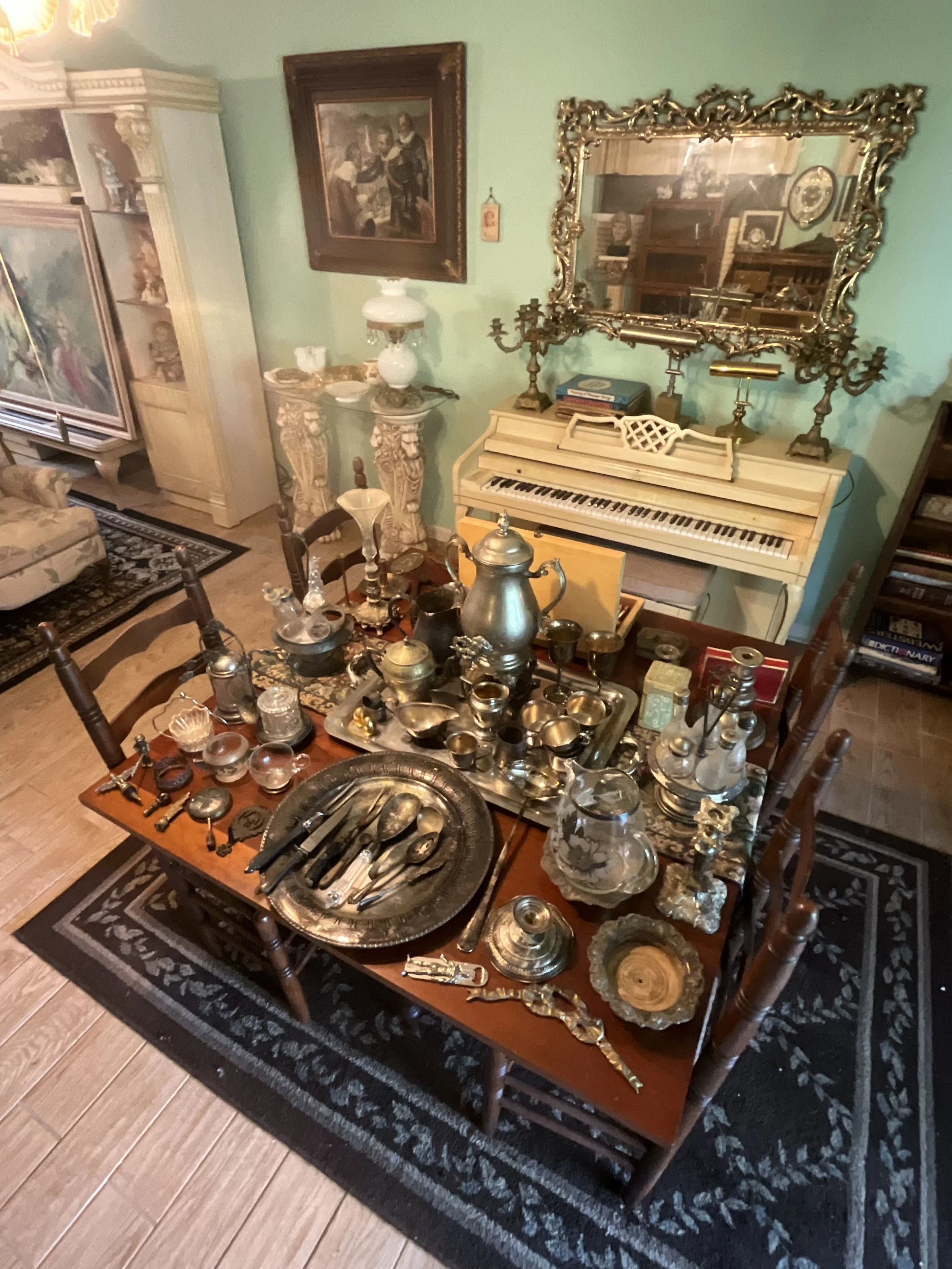 A dining room with a table filled with antique silverware and collectibles, a white upright piano with a gilded mirror above it, and paintings and a cabinet in the background.