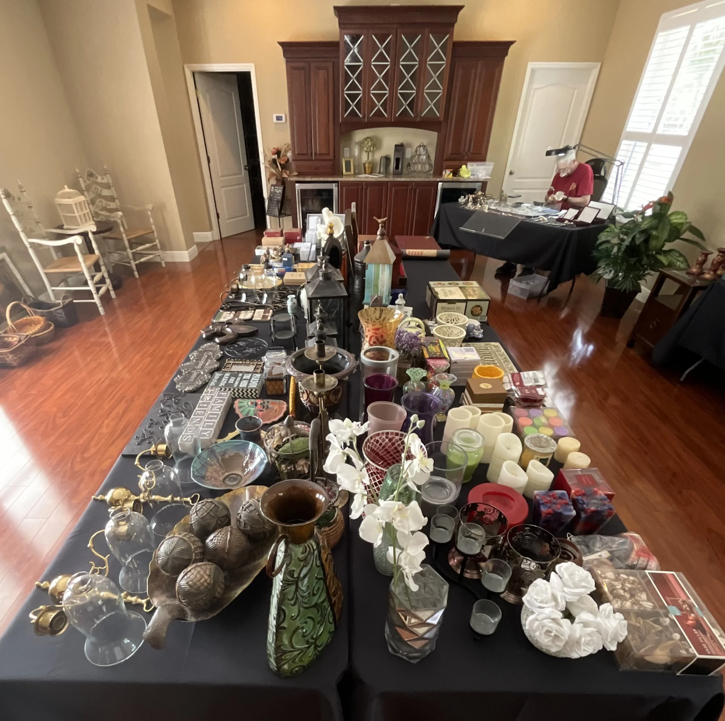 A room with a large black table filled with various collectibles, decorations, and small items, with a wooden cabinet against the wall and other furniture, including chairs and side tables, in the background.