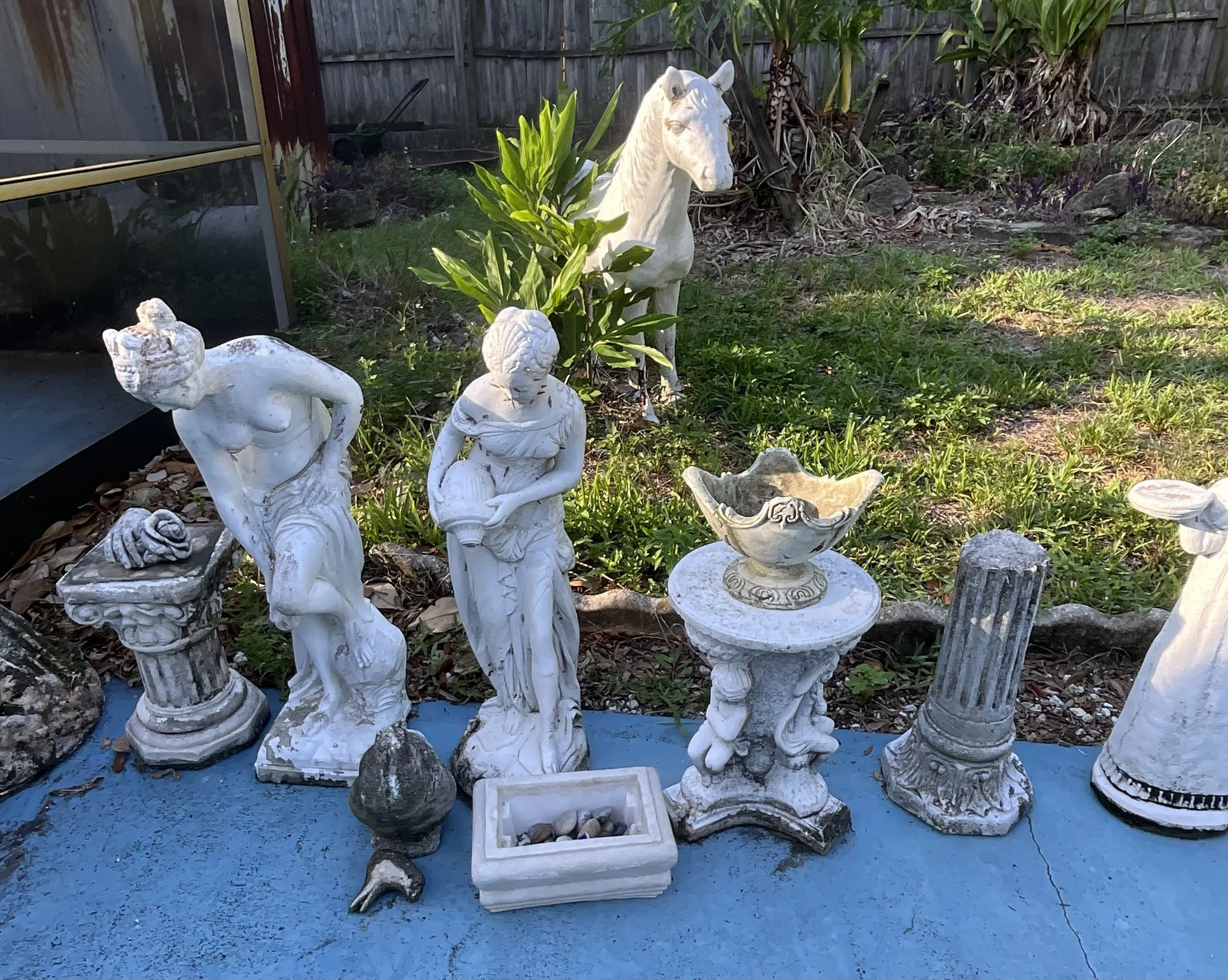 Collection of white classical-style garden statues and ornaments. Including a lounging female figure, a standing female figure, a bowl on a decorative pedestal, and a small bowl with rocks, set on a blue surface in an outdoor garden.