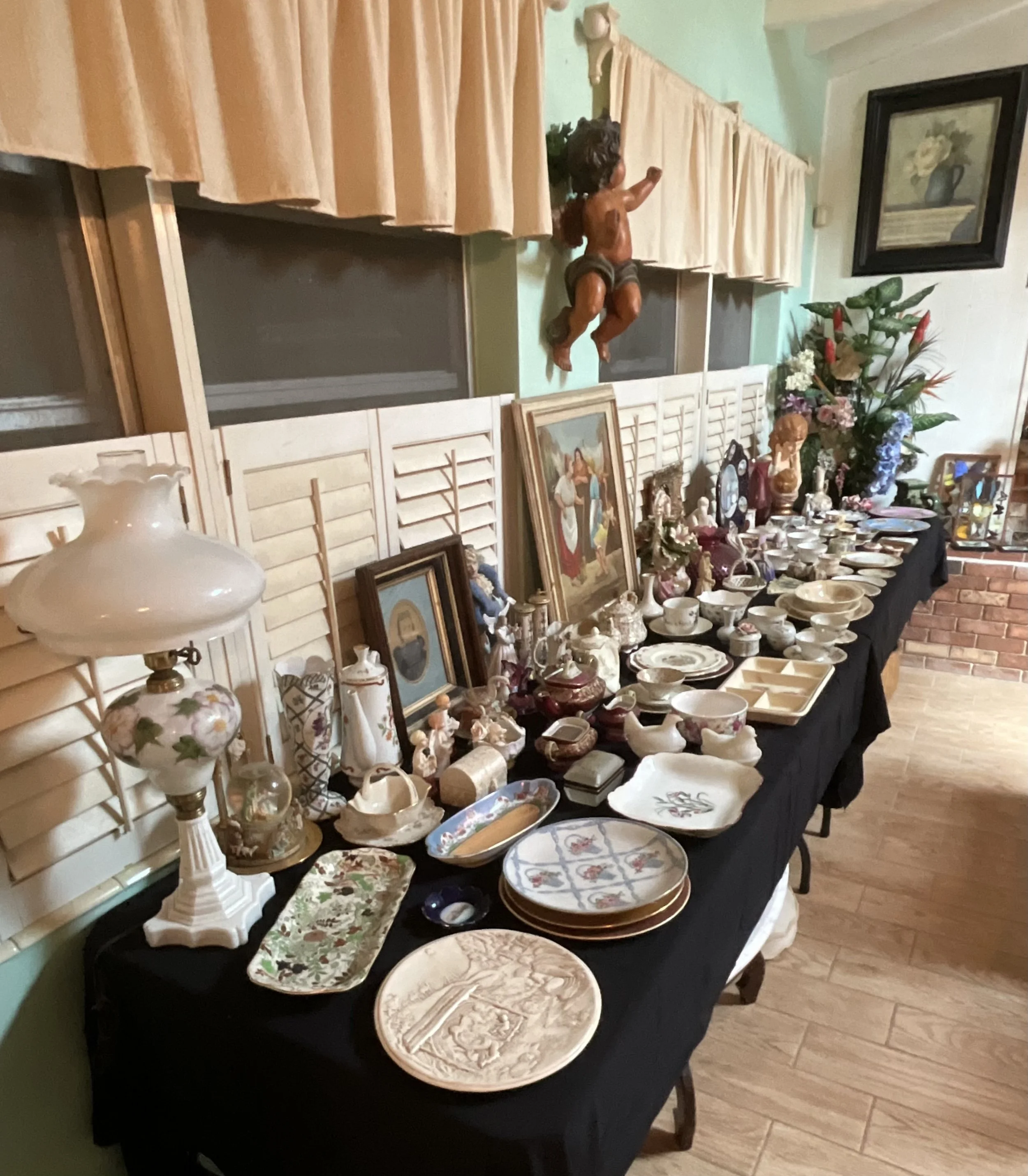 A long table covered with a black cloth displaying various vintage and decorative china, pottery, and artwork in a room with green walls, a brick fireplace, and wall-mounted decorations.