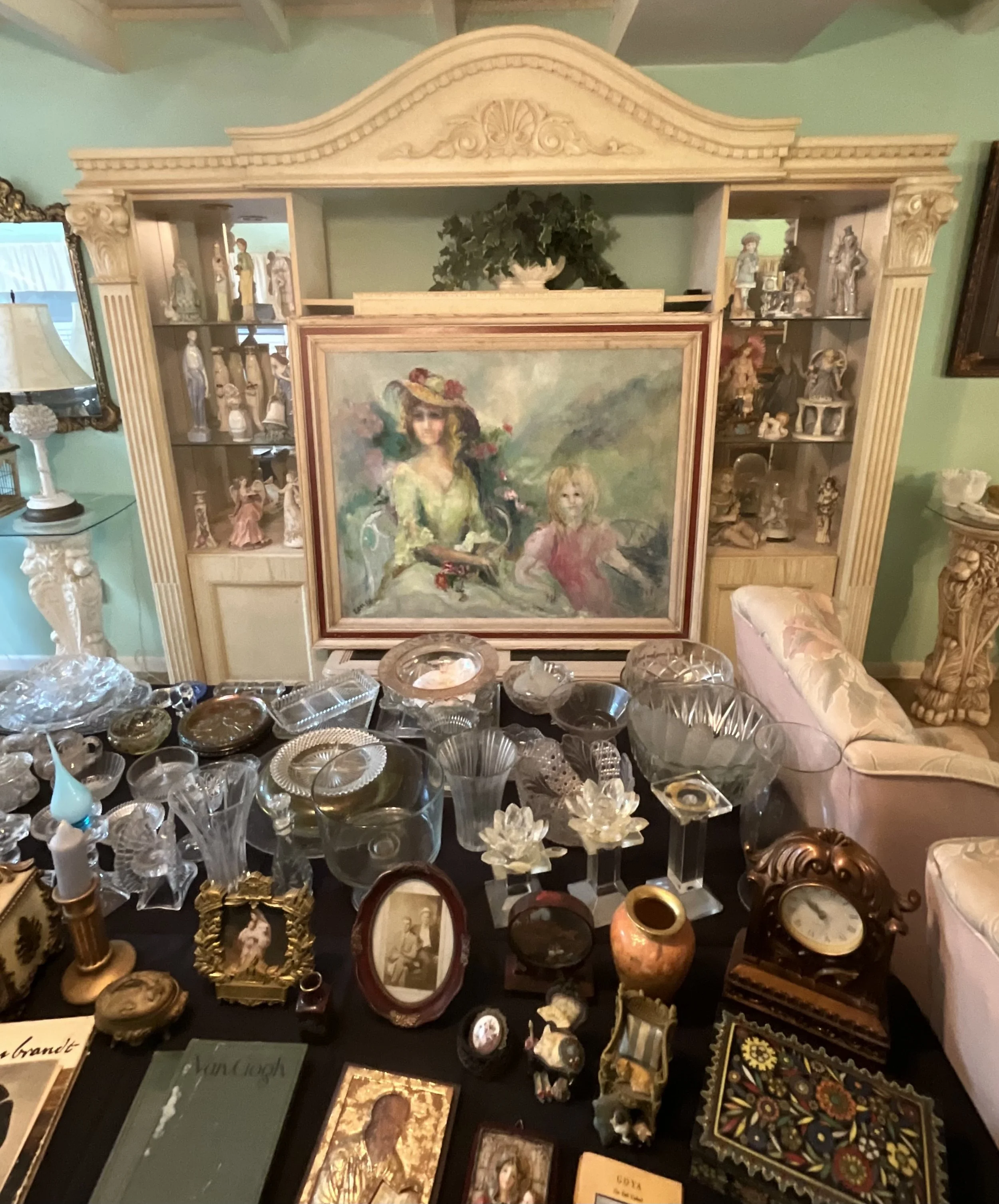 A room with a large, ornate cream-colored shelf containing figurines. In front are various glass and ceramic items on a black table, with a painting of a woman and a child on the shelf behind.
