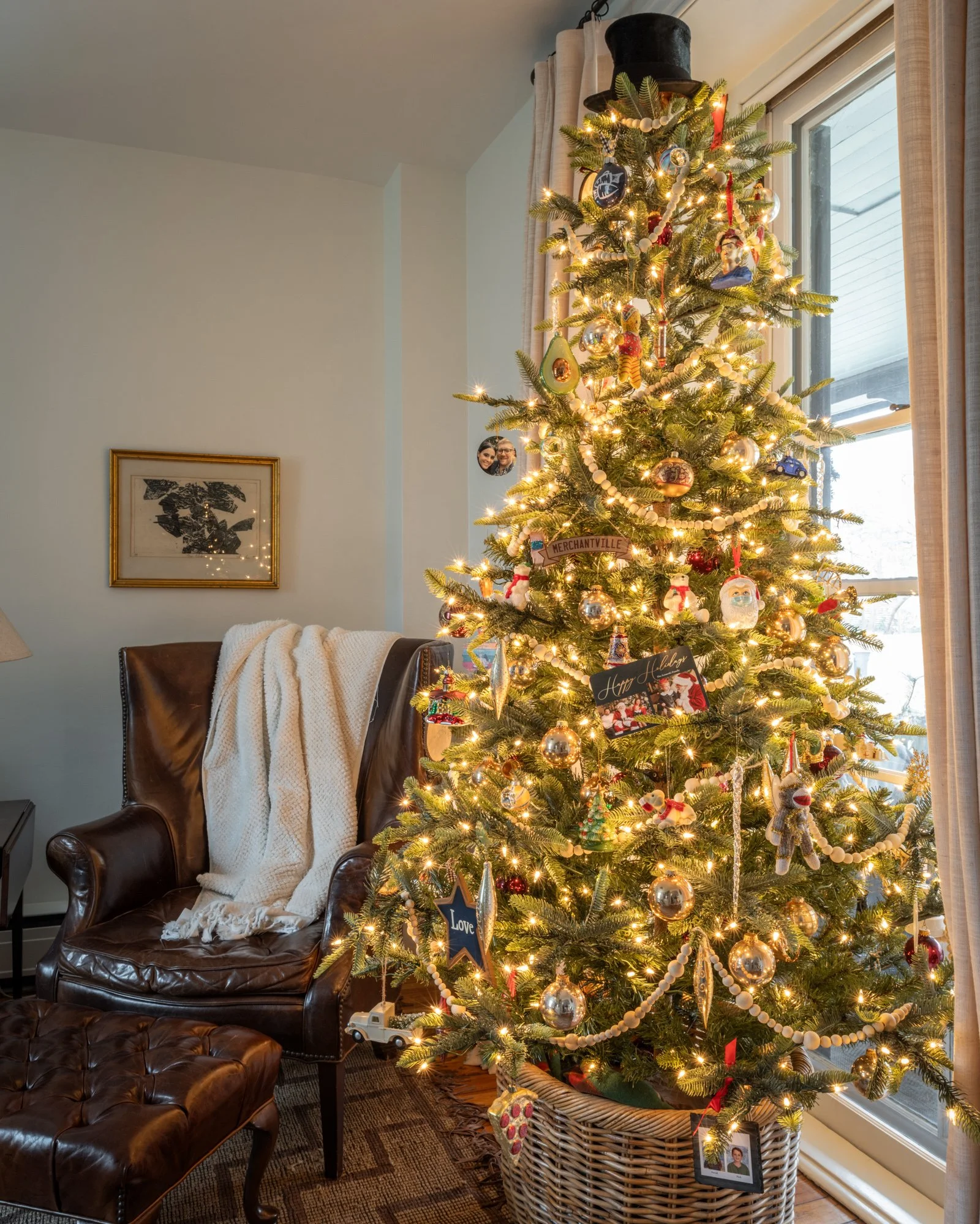 Decorated Christmas tree with lights and ornaments in a cozy living room, next to a leather armchair with a blanket, window with curtains in the background, and framed picture on wall.