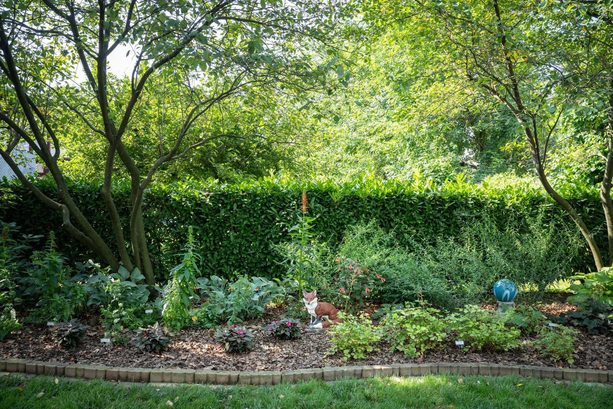 Garden with lush green trees and bushes, a small fox statue, a globe, and flowering plants, all on a landscaped area with a brick-edged border.