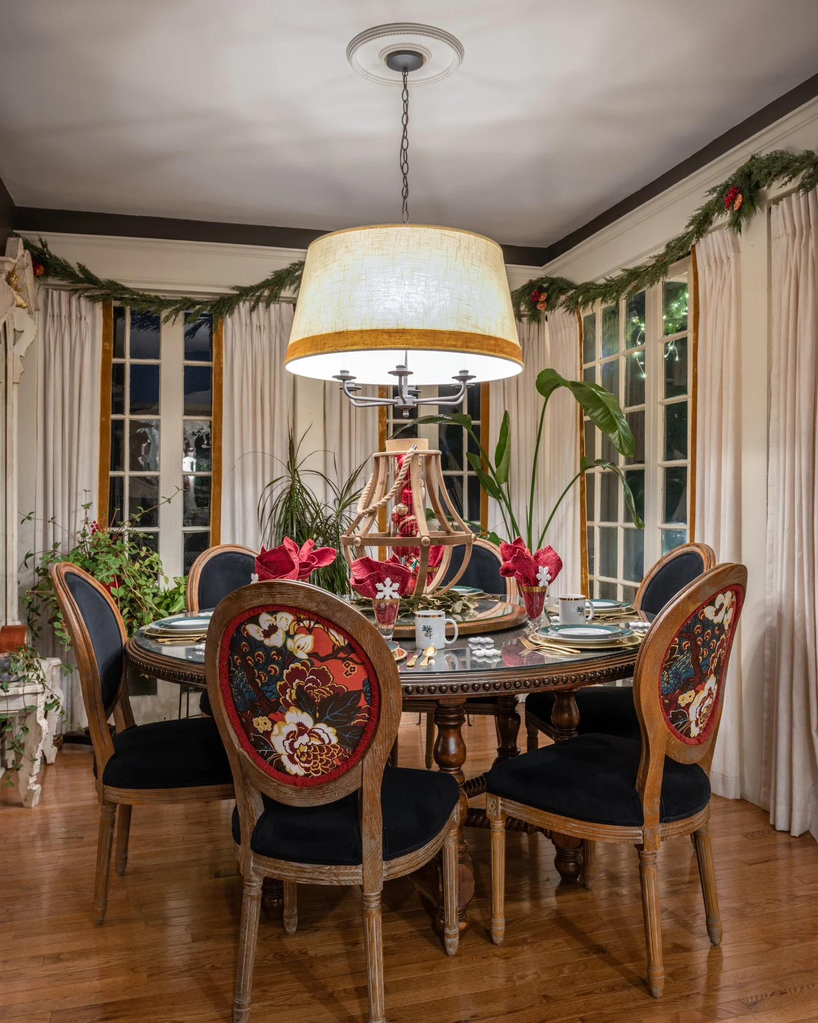 A dining room with a round wooden table surrounded by eight chairs with black cushioned seats and floral upholstery. The table is decorated with red poinsettias and holiday-themed items, and there is a large chandelier with a beige lampshade hanging 