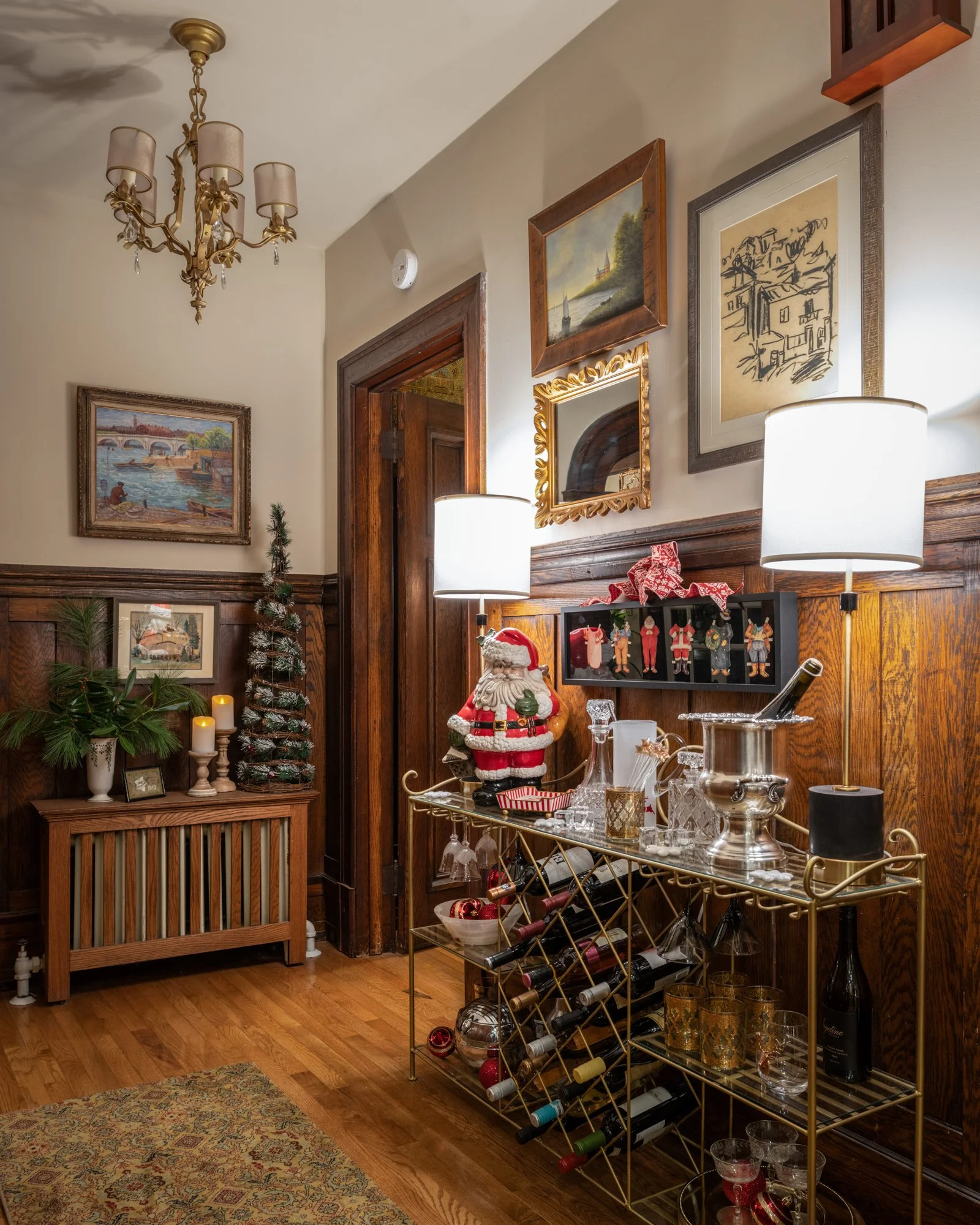 A cozy, decorated corner of a home with a wooden floor, a small Christmas tree, paintings on the wall, a chandelier, table lamps, and a gold wine rack filled with wine bottles and glasses, with festive decorations and candles.