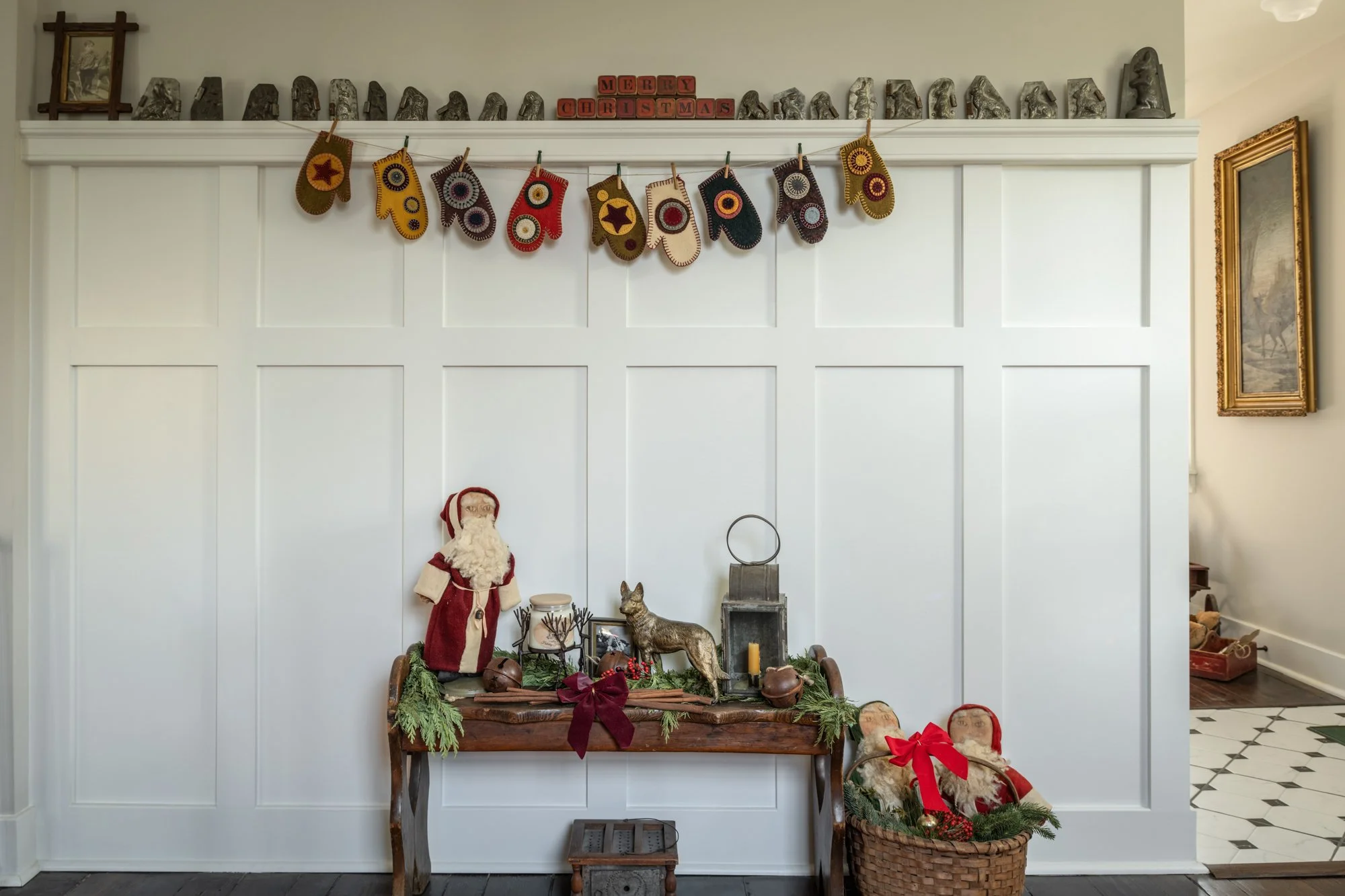 A Christmas mantle decoration scene with stuffed Santa and reindeer figures, candles, pine branches, and a woven basket with holiday decor, set against white wall paneling with a 