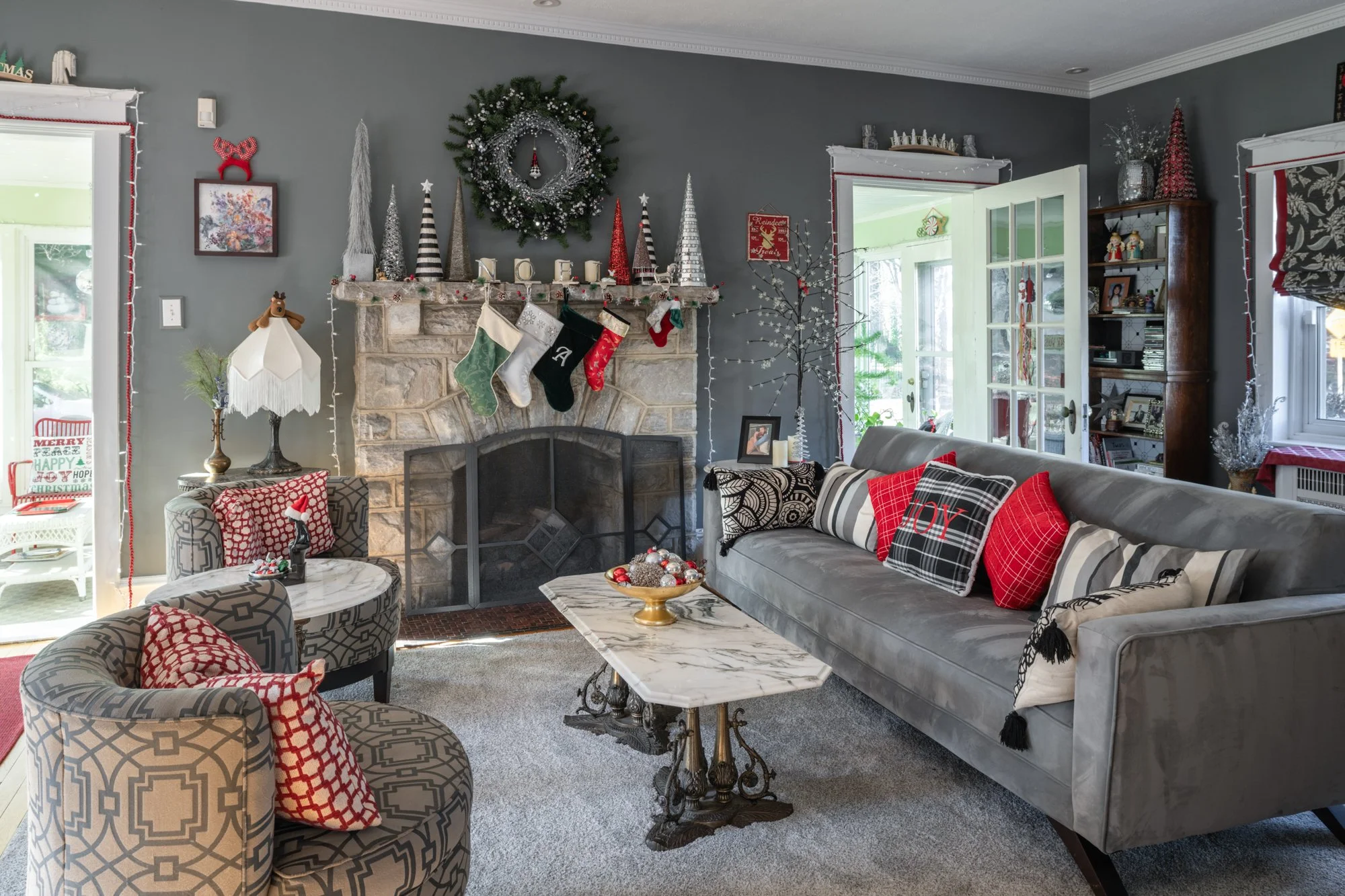 Living room decorated for Christmas with a stone fireplace, Christmas stockings, wreath, cone-shaped holiday decorations, and a sofa with red and black pillows, with a marble coffee table and a patterned armchair.
