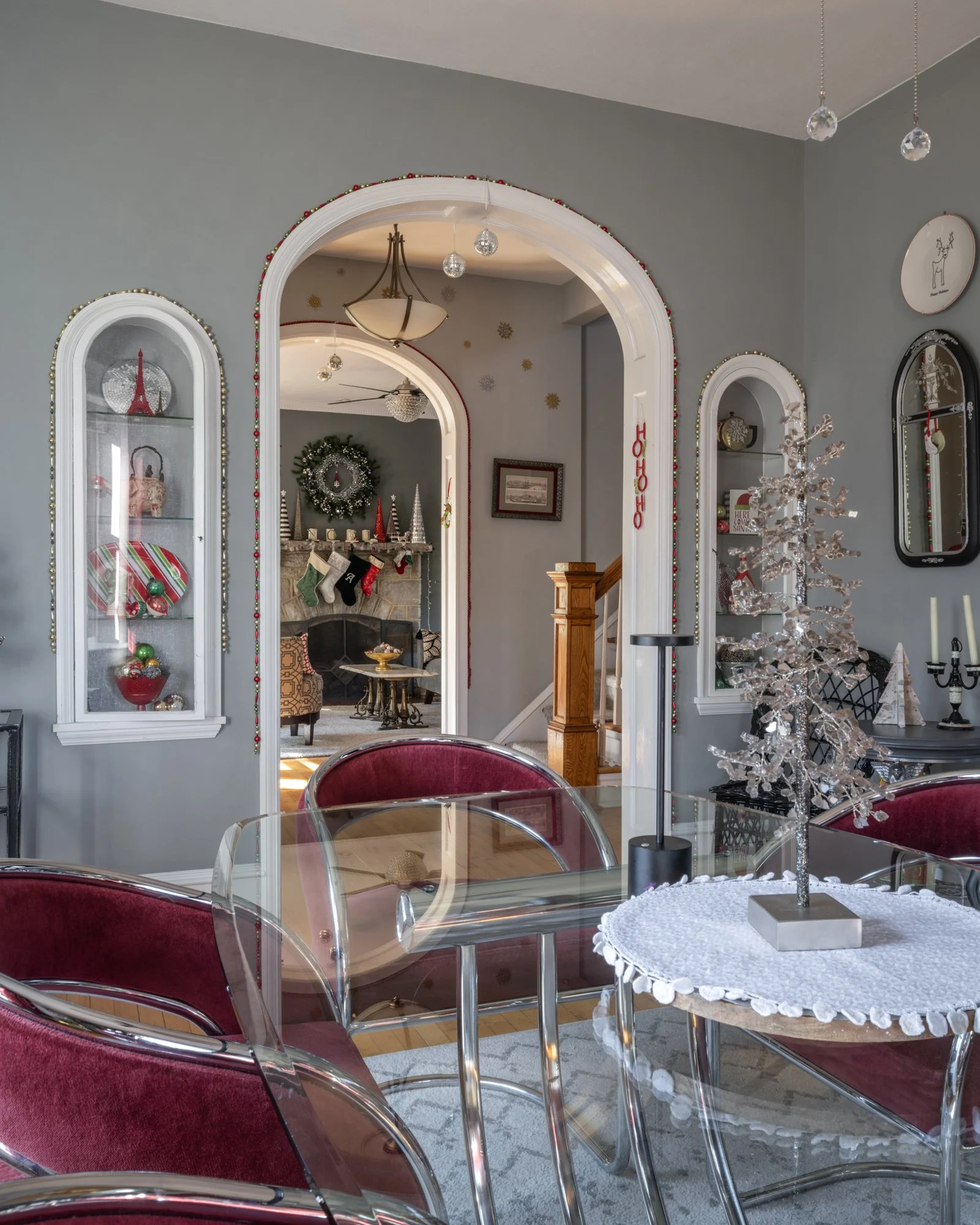 Interior of a home decorated for Christmas with a glass-top table and red velvet chairs, a small white Christmas tree, and a fireplace with stockings and holiday decor.